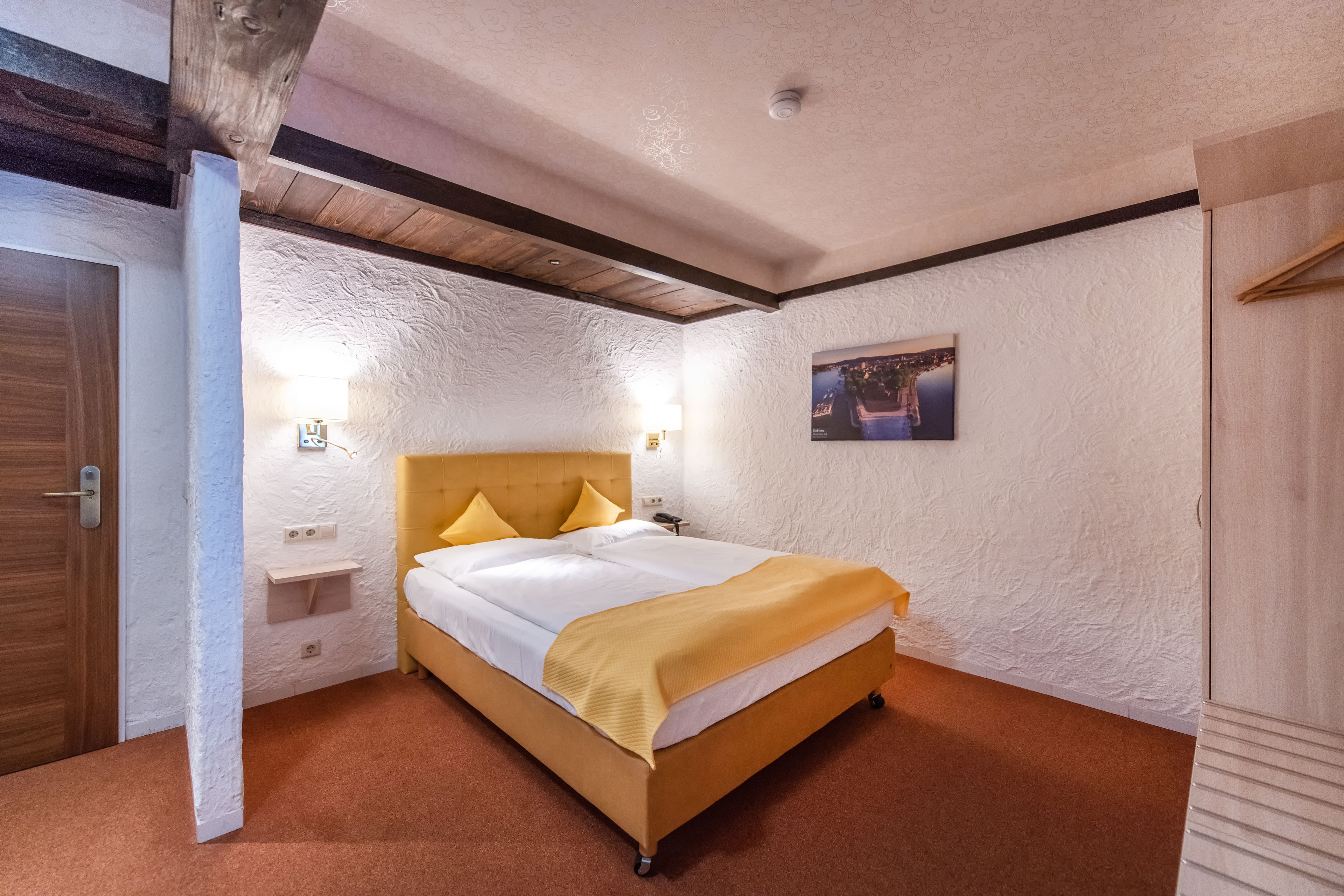 Quarto com cama de madeira, paredes brancas com textura, teto de madeira e uma pequena mesa de cabeceira com abajur.