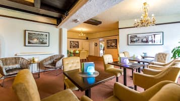 Um hall de hotel com uma mistura de mobiliário moderno e clássico, incluindo sofás e poltronas, uma mesa de centro com abajur, e quadros nas paredes.