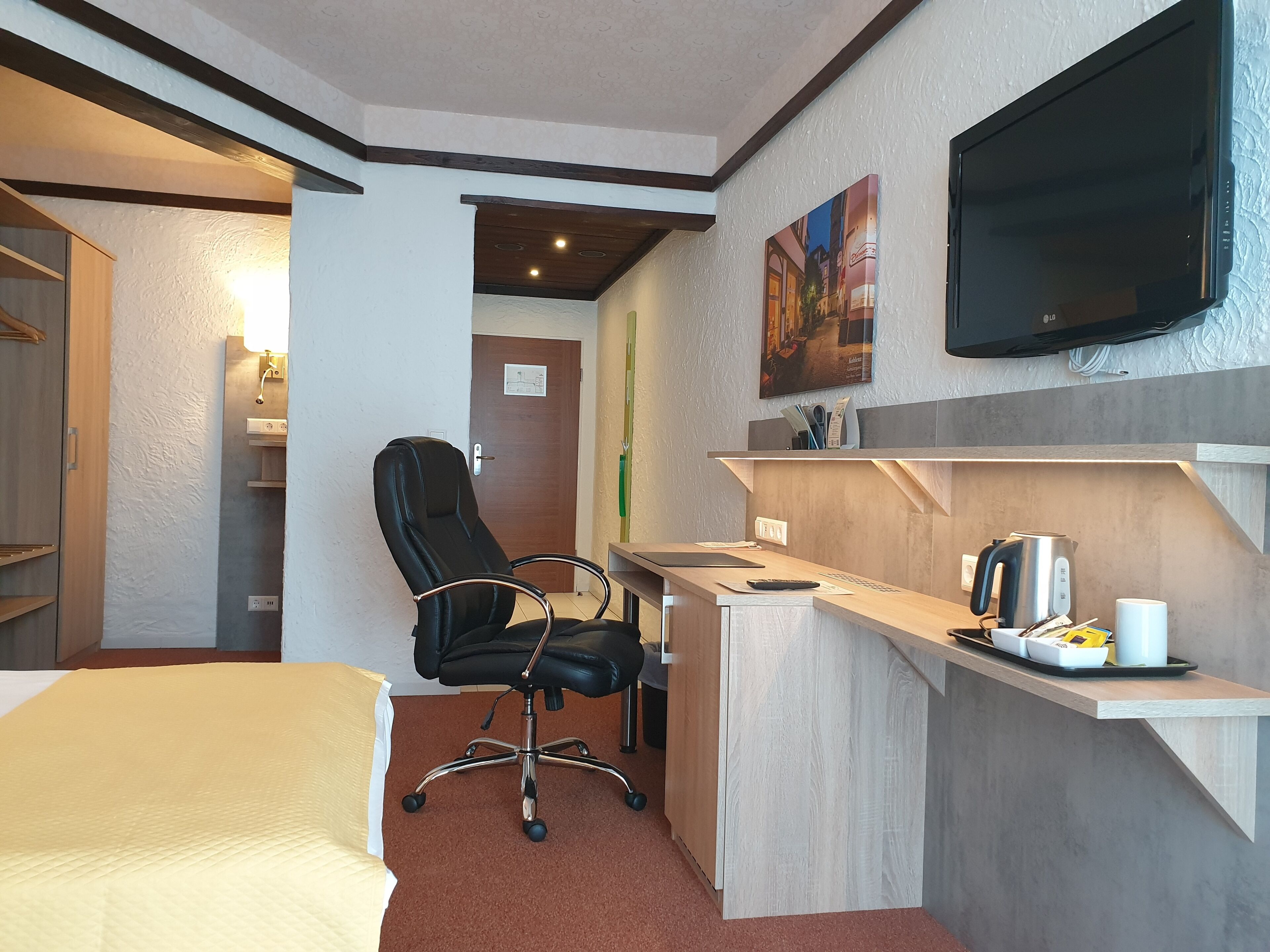 Habitación de hotel con cama, escritorio con hervidor, silla y televisor montado en la pared.