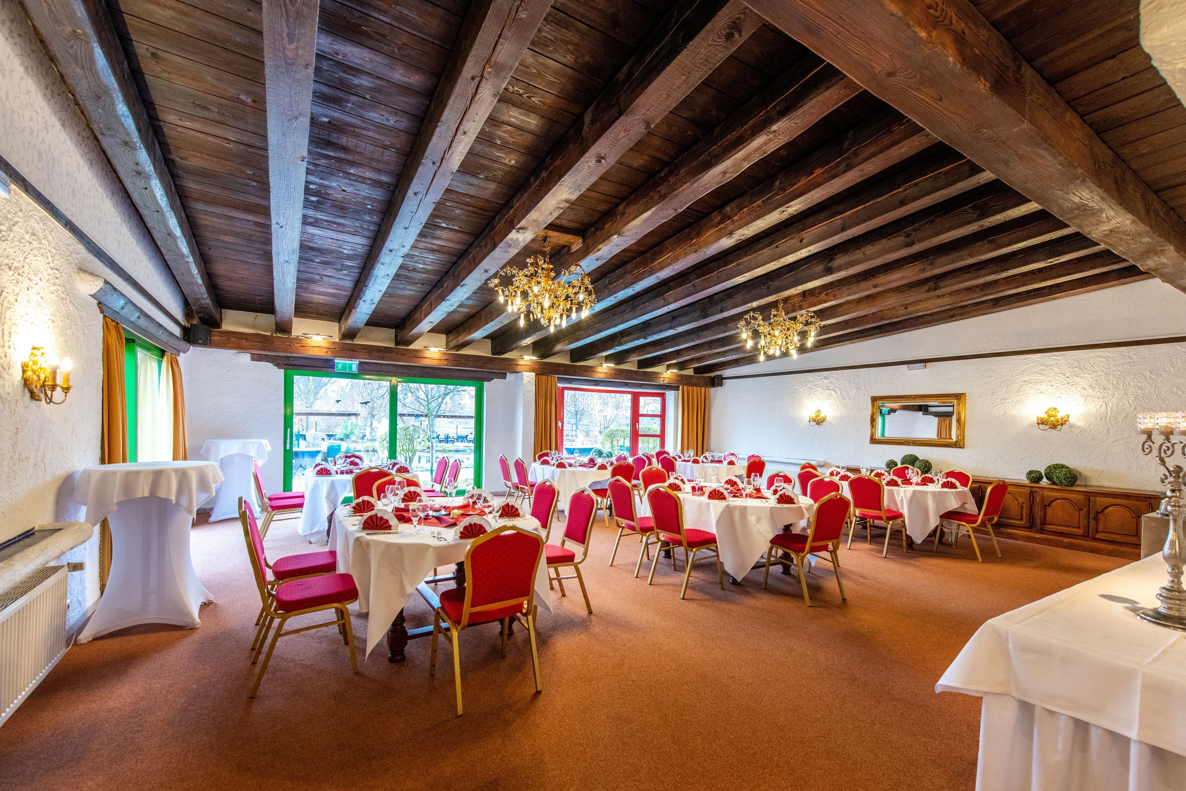 Un salón de banquetes con vigas de madera, mesas blancas, sillas rojas y una araña.