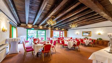 Un salón de banquetes con vigas de madera, mesas blancas, sillas rojas y una araña.