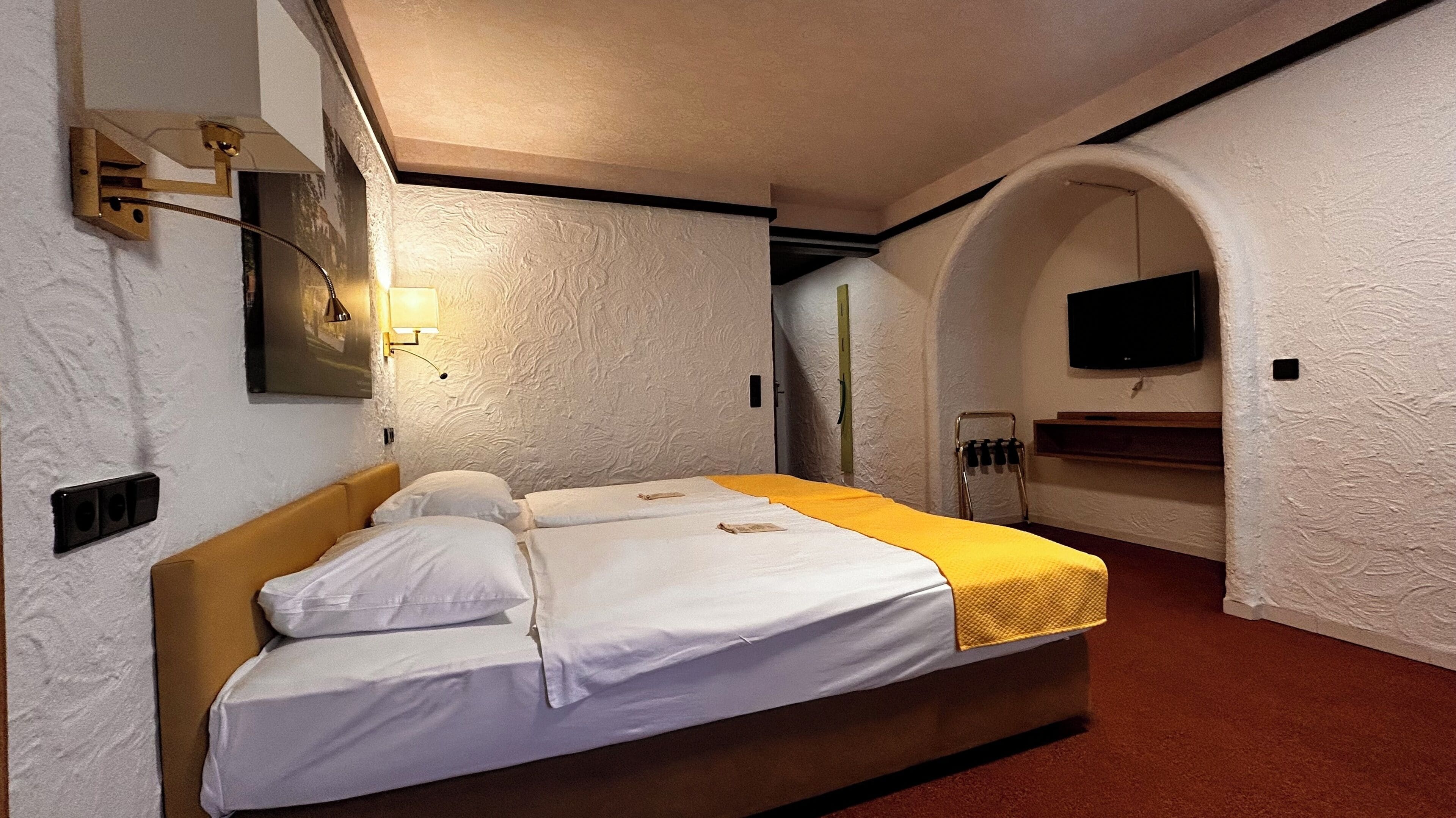 Une chambre d’hôtel avec un lit, un téléviseur fixé au mur et un petit coin salon avec un banc.