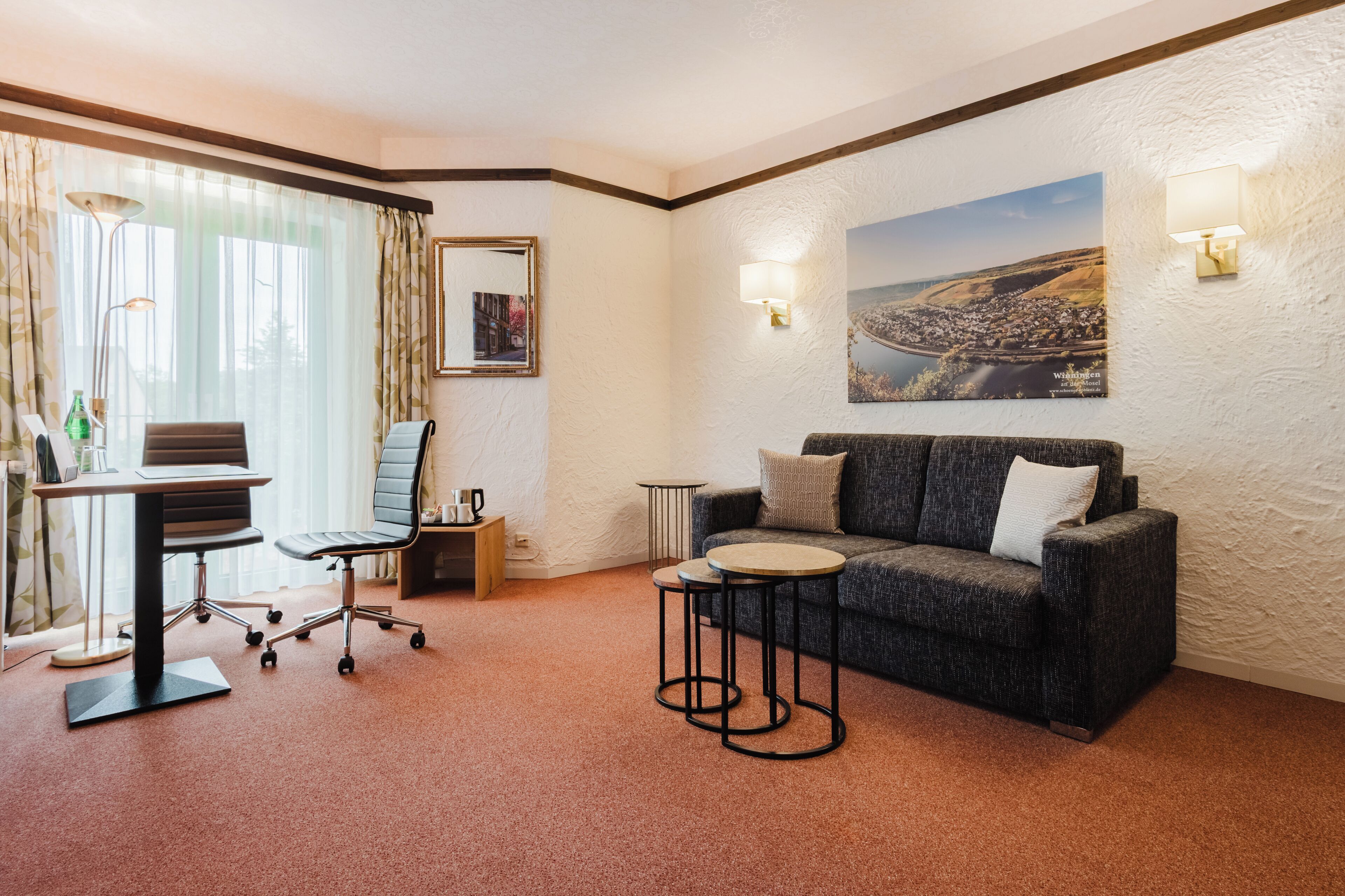 Habitación de hotel con escritorio, silla de oficina, sofá, dos mesitas auxiliares y un ventanal con cortinas.
