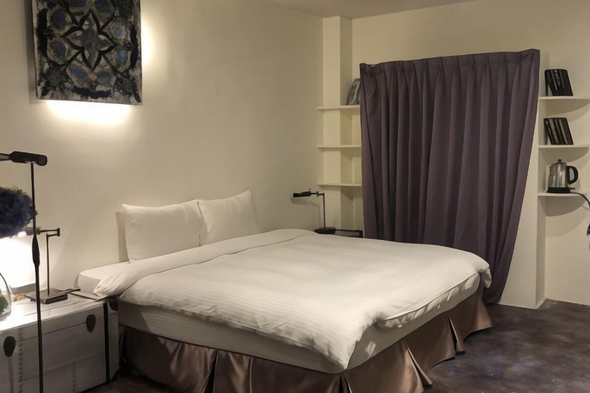 A bedroom with a bed, bedside tables, a lamp, a shelf with books, and a curtain.