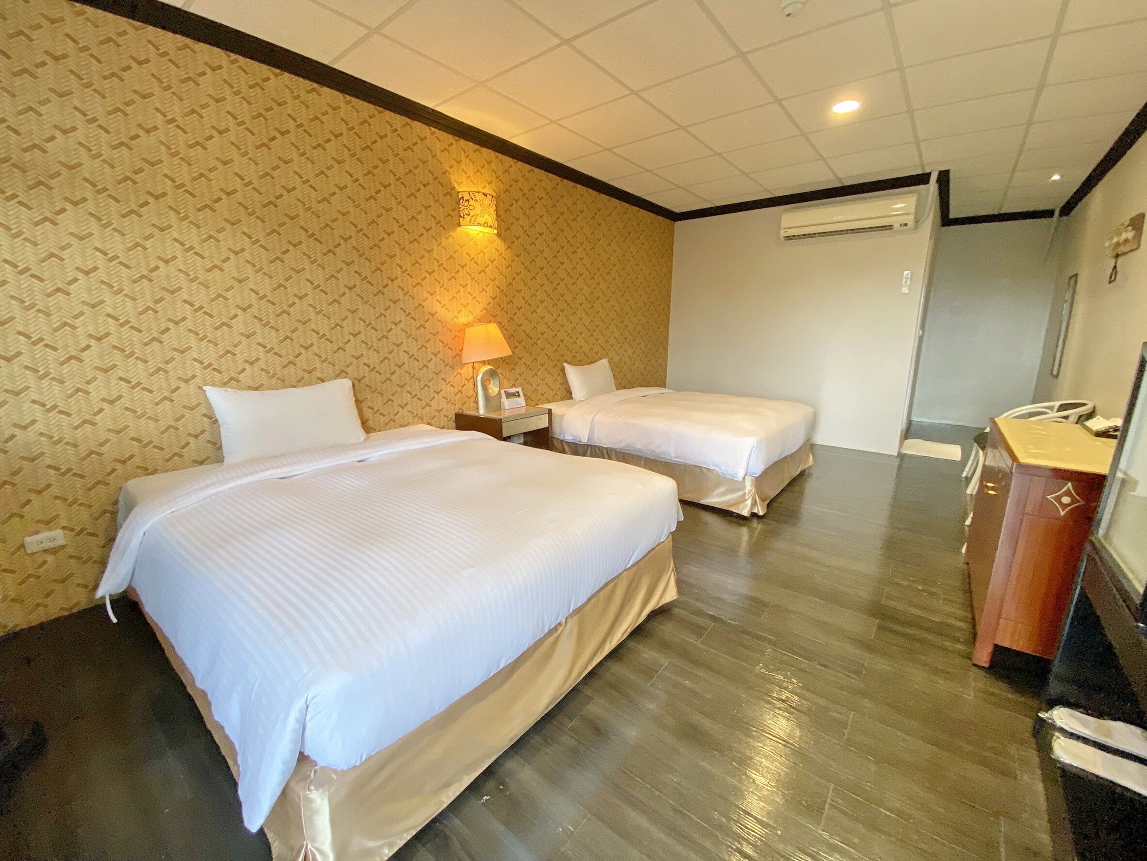 A hotel room with two beds, a nightstand, a lamp, an air conditioning unit, and a wooden desk with a telephone.