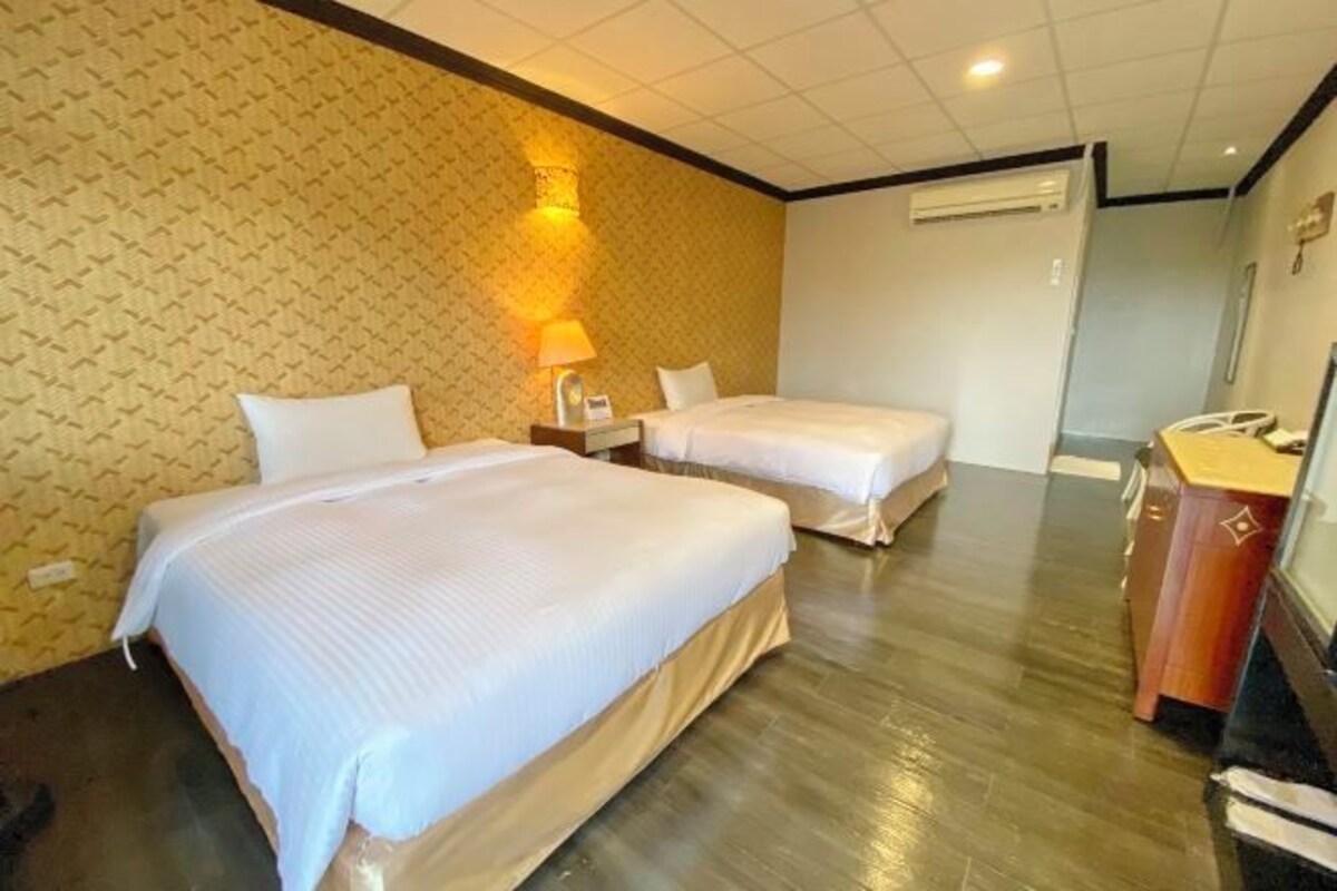 A hotel room with two beds, a bedside table with a lamp, a wooden dresser, and a wall-mounted air conditioner.