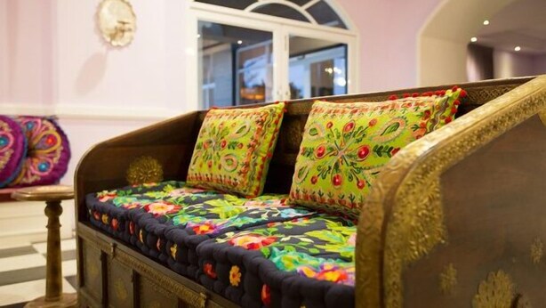 A sofa with colorful floral upholstery and decorative pillows, set against a pink wall with a decorative mirror and a window in the background.