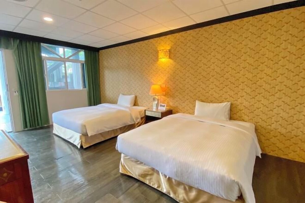 Une chambre d’hôtel avec deux lits, une table de chevet, une lampe et une fenêtre avec des rideaux verts.