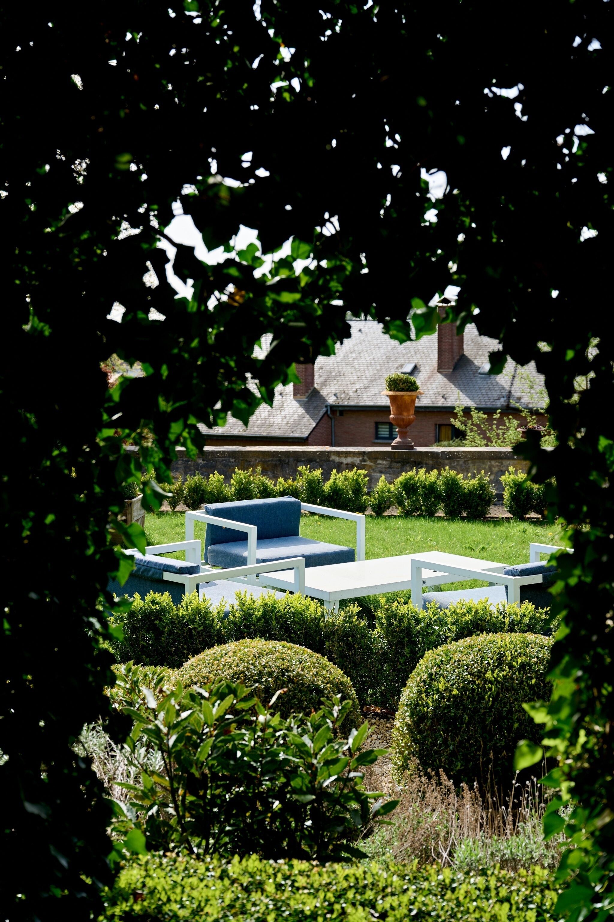 Un jardín con setos bien cuidados, una zona de estar exterior blanca y un edificio de ladrillo con una chimenea al fondo.