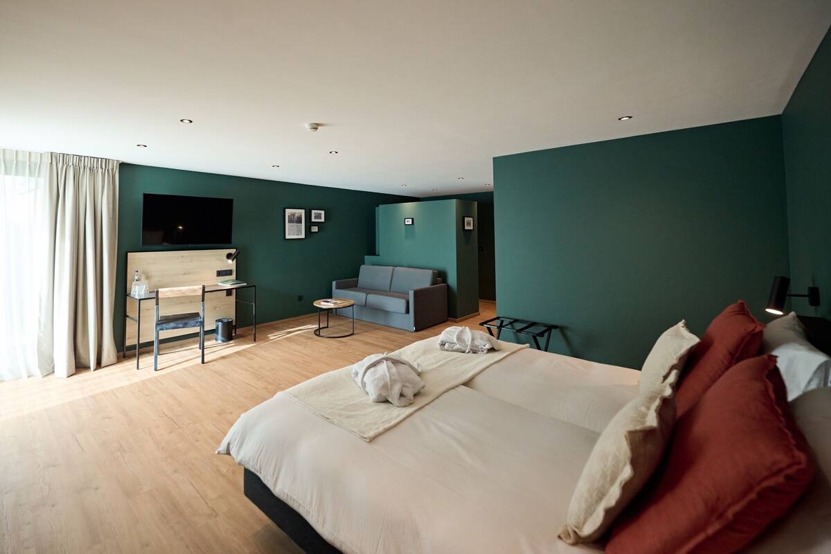 A modern hotel room with a large bed, a desk, a sofa, and a TV mounted on the wall.