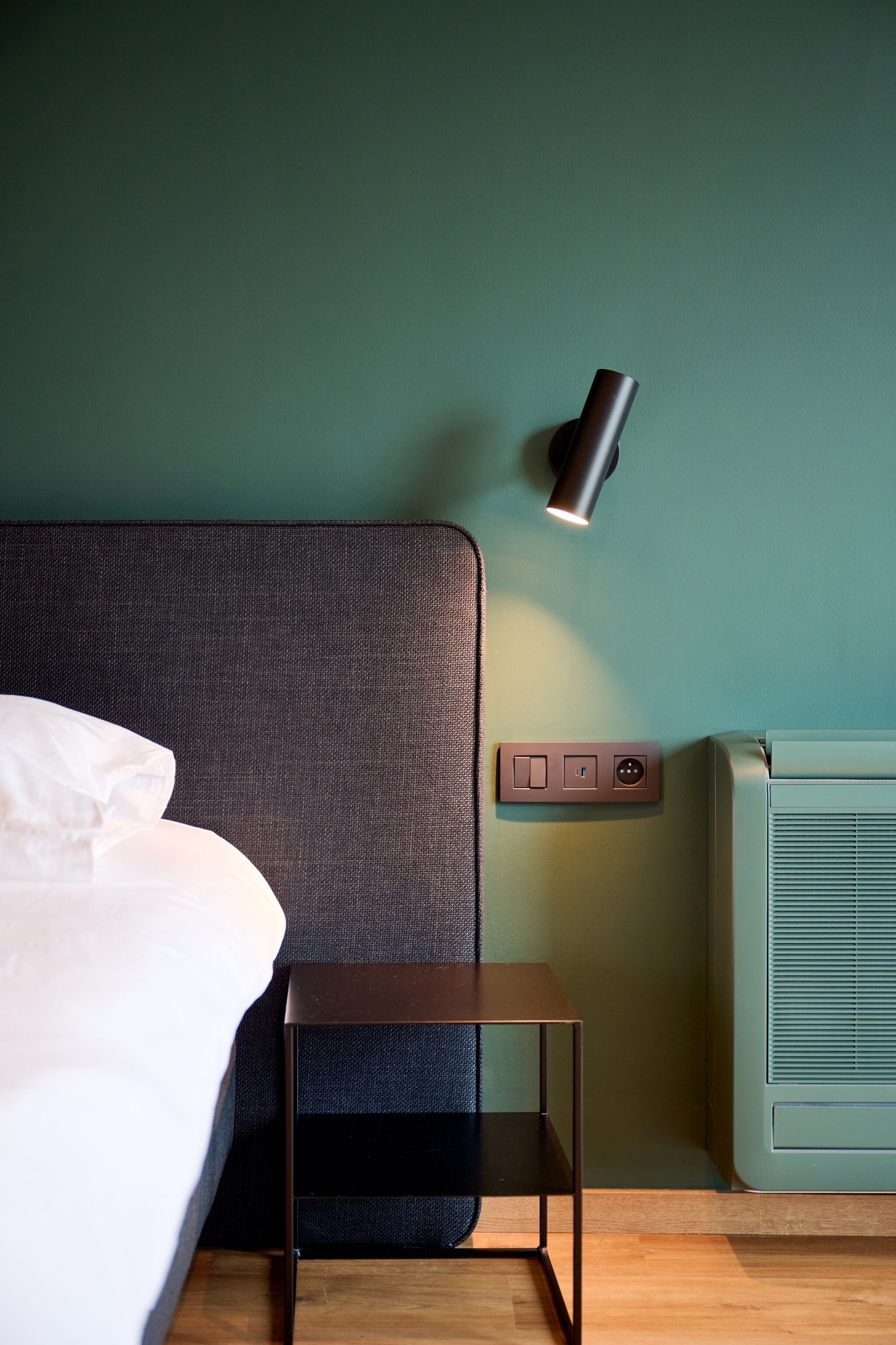 Una cama bien hecha con un cabecero oscuro, una mesita de noche pequeña, una lámpara de pared y un enchufe.