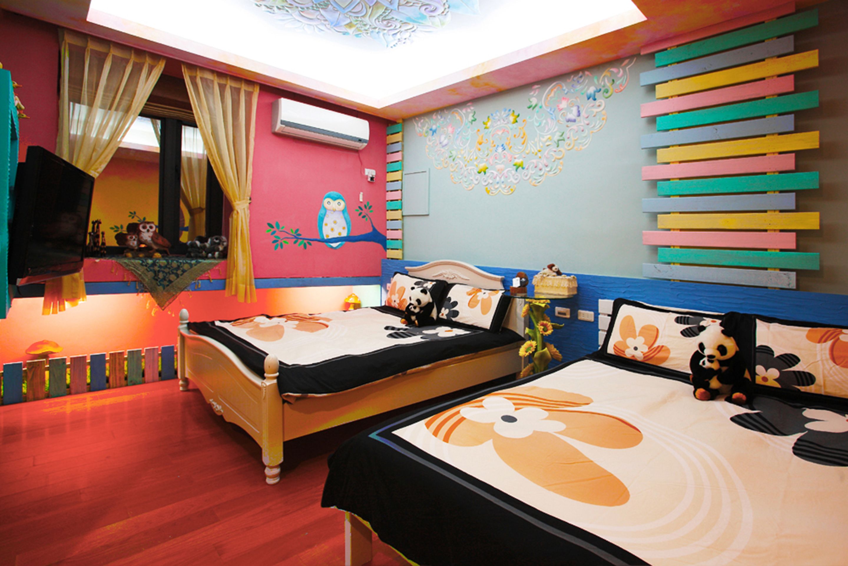 A room with two beds, a window with curtains, a wall with colorful decorations, and a ceiling light.
