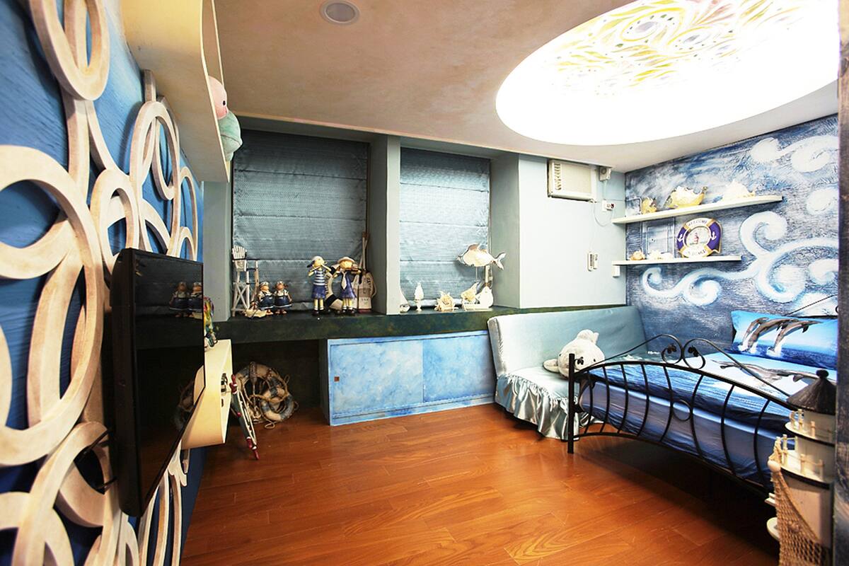 A room with a bed, a desk, and a chair. There are shelves with decorative items and a window with blinds.