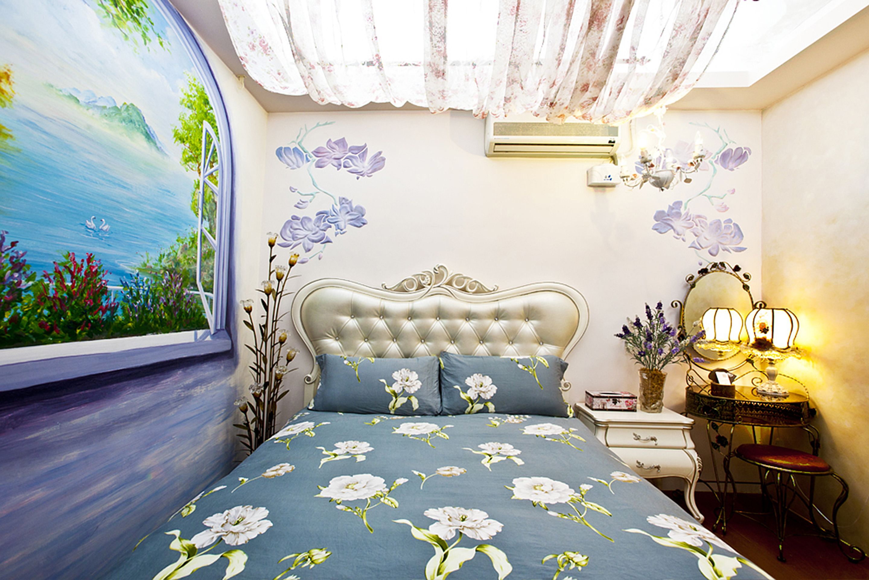 Quarto com colcha florida, cabeceira estofada, penteadeira com espelho e um mural na parede.