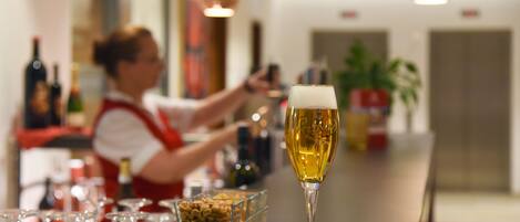 A bar with a glass of beer, a bartender, and a glass bowl of nuts on the counter.