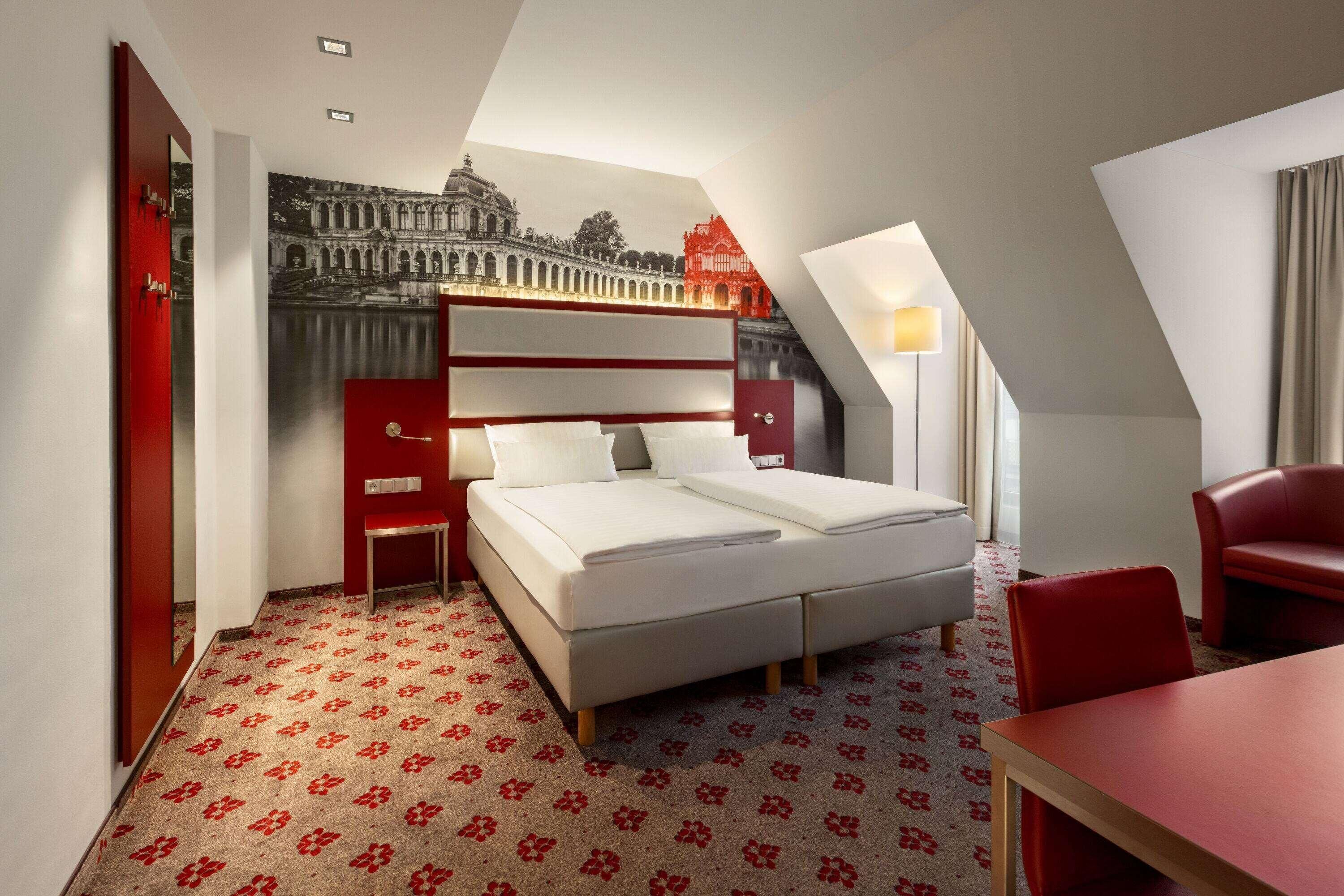 A hotel room with a bed, a desk, a chair, a red wardrobe, and a large window.