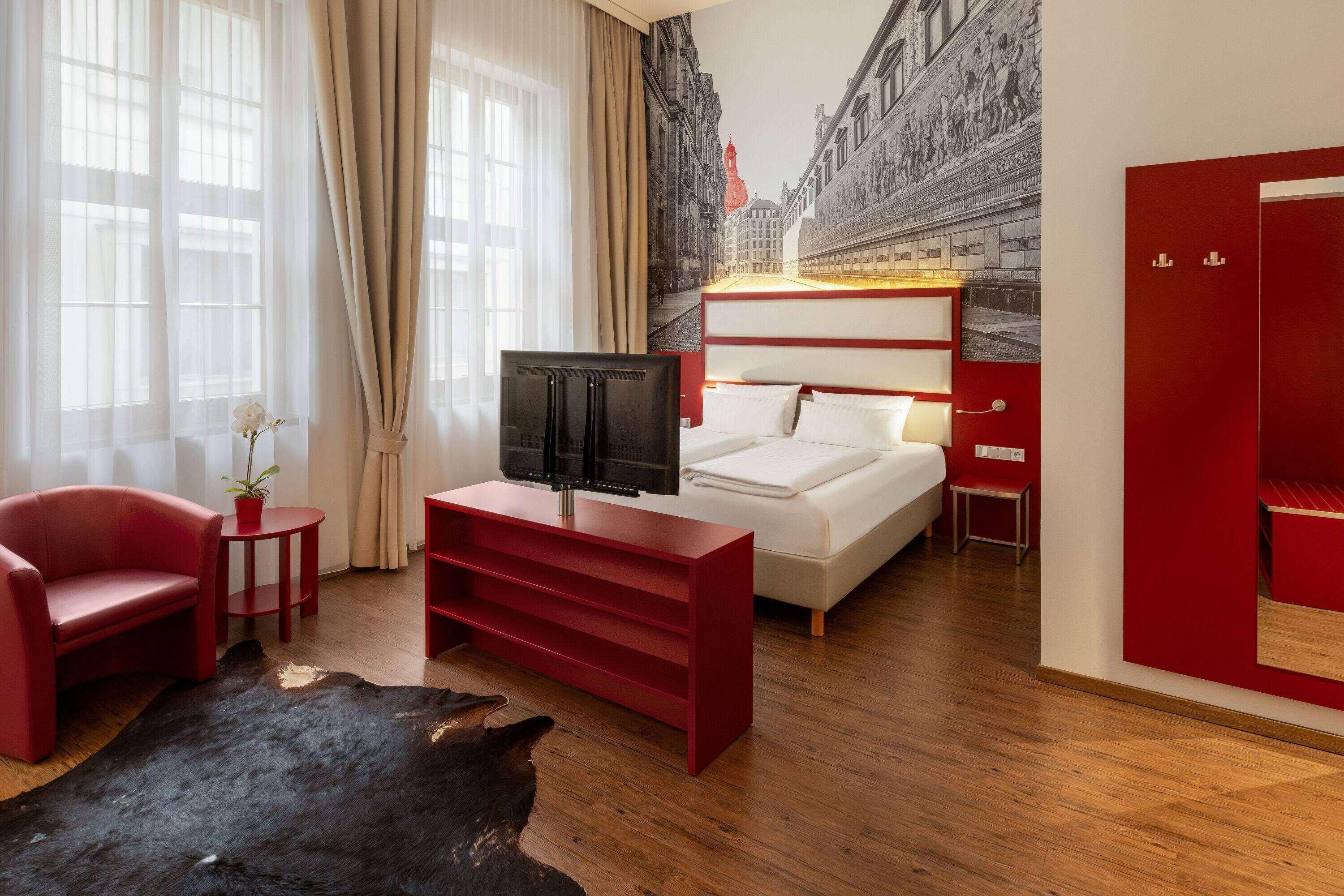 A hotel room with a bed, a TV, a red sofa, a small table, and a red cabinet.