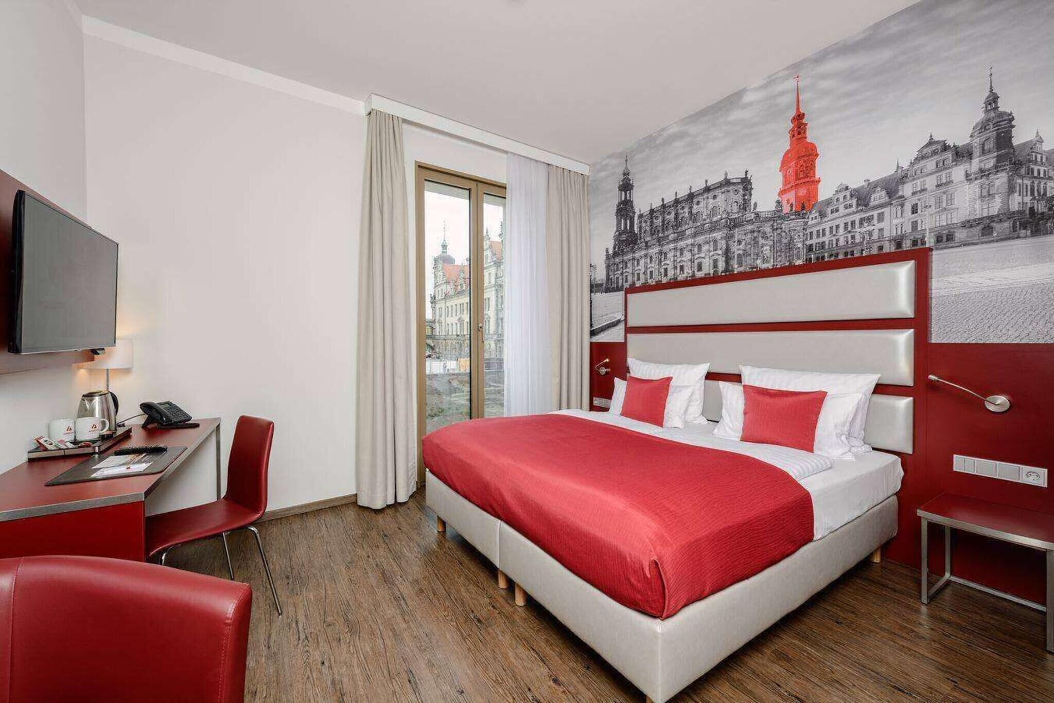 A hotel room with a large bed, a desk with a chair, a television, and a cityscape mural on the wall.