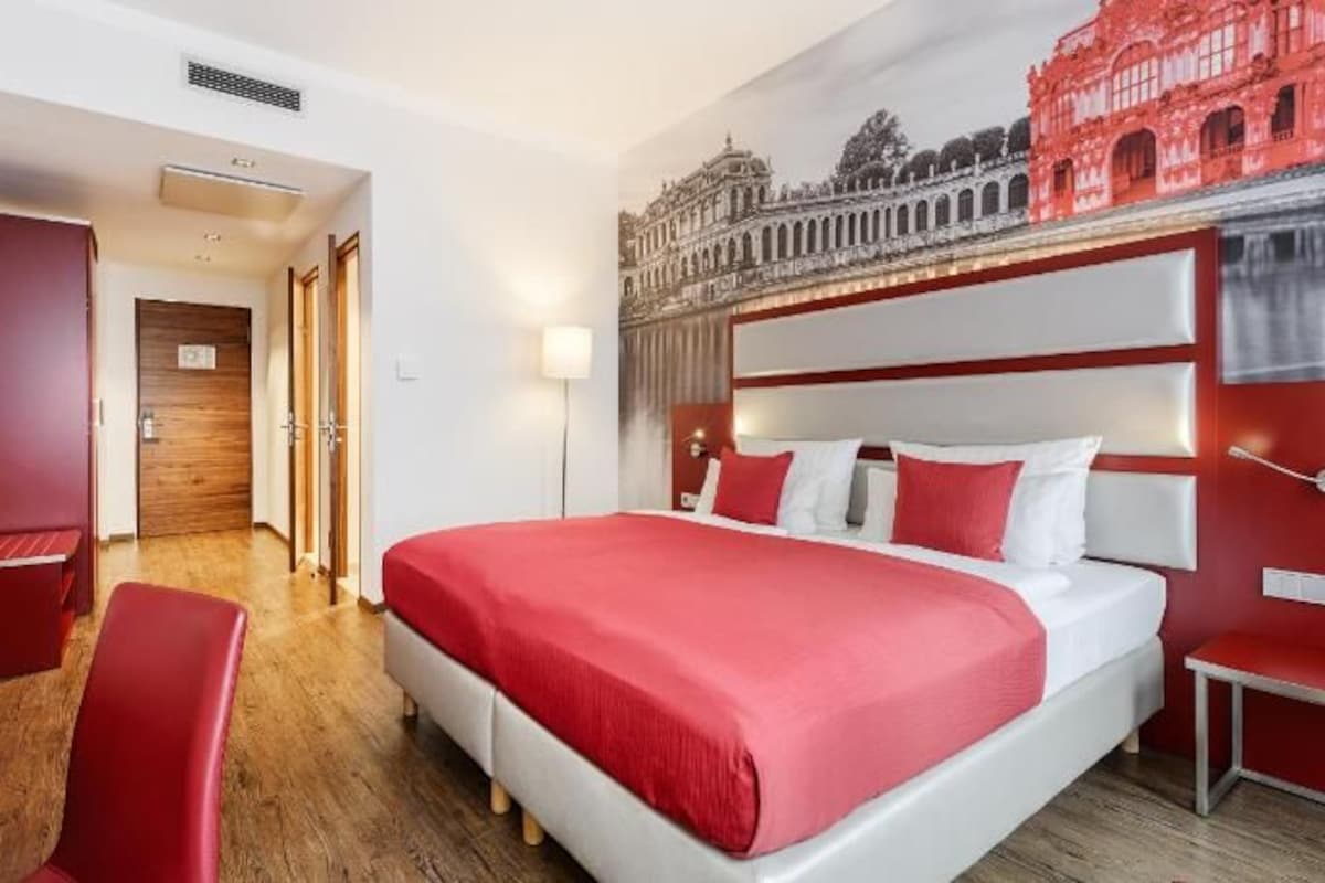 A hotel room with a large bed, a desk, a chair, and a view of a building through the window.