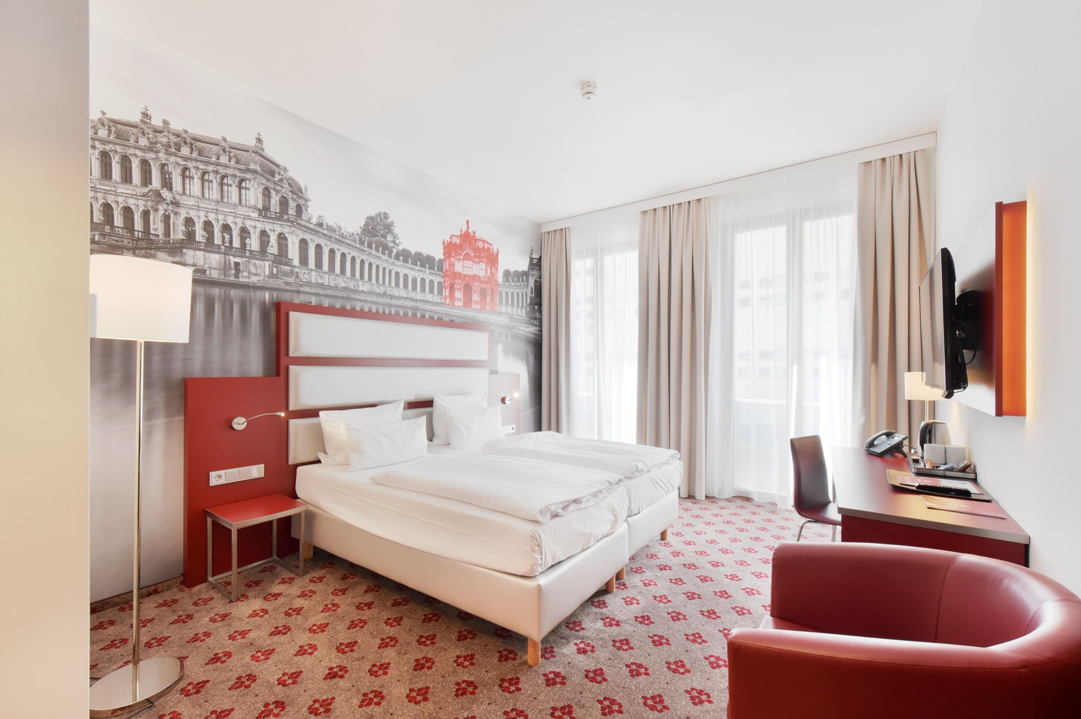 Ein Hotelzimmer mit einem großen Bett, einem Schreibtisch mit Telefon, einem roten Sessel und einem an der Wand montierten Fernseher.