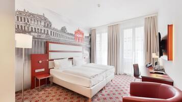 Ein Hotelzimmer mit einem großen Bett, einem Schreibtisch mit Telefon, einem roten Sessel und einem an der Wand montierten Fernseher.