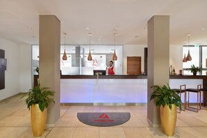Uma área de recepção com design moderno, incluindo um balcão de atendimento, plantas em vasos e luminárias pendentes.