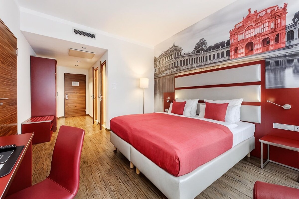 A hotel room with a large bed, red and white decor, a wooden floor, and a mural of a historical building.