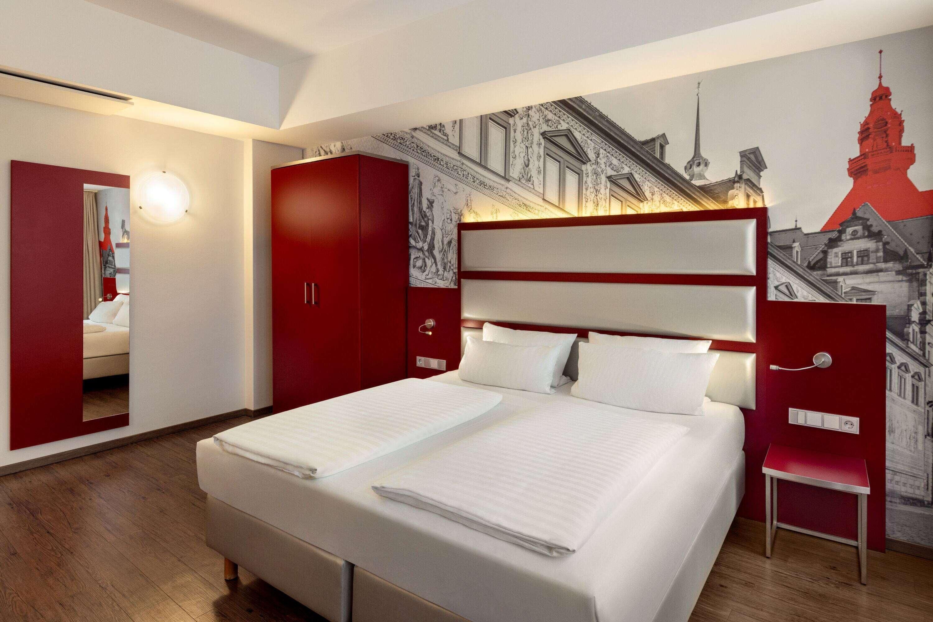 A modern hotel room with a large bed, a red wardrobe, a mirror, and a cityscape mural on the wall.