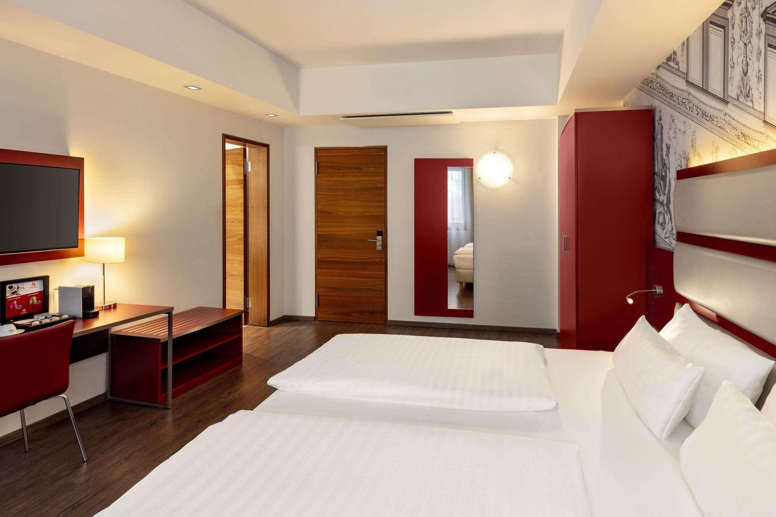 A hotel room with a bed, a desk, a TV, and a red accent wall.