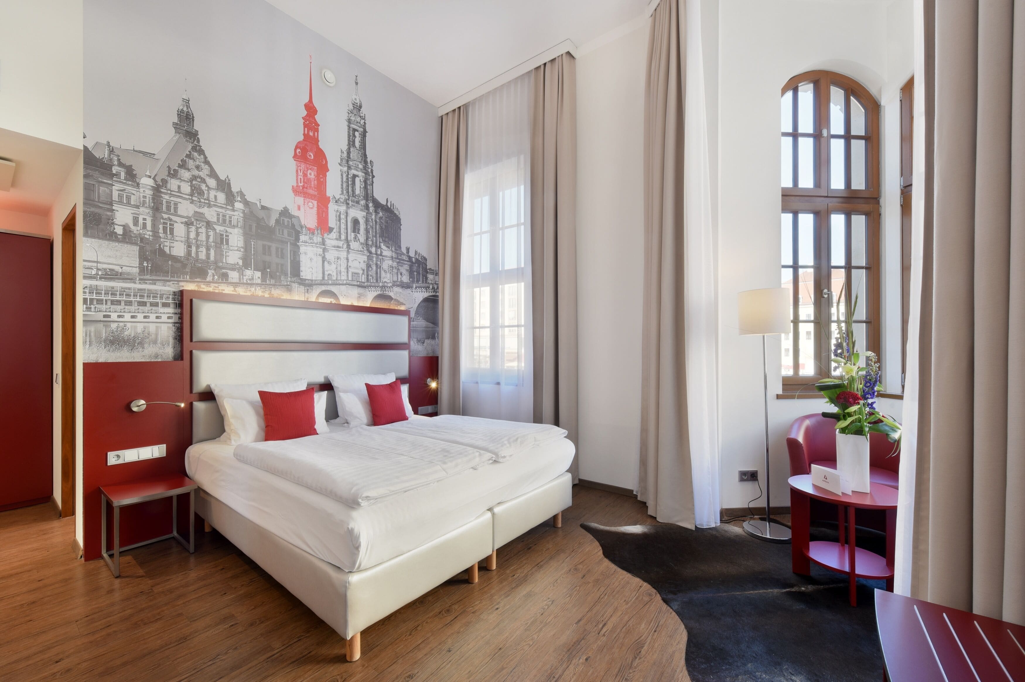 A hotel room with a large bed, a desk, a chair, a small table with a vase of flowers, and a wall mural.