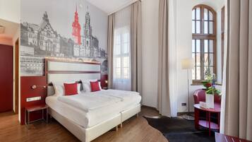 A hotel room with a large bed, a desk, a chair, a small table with a vase of flowers, and a wall mural.