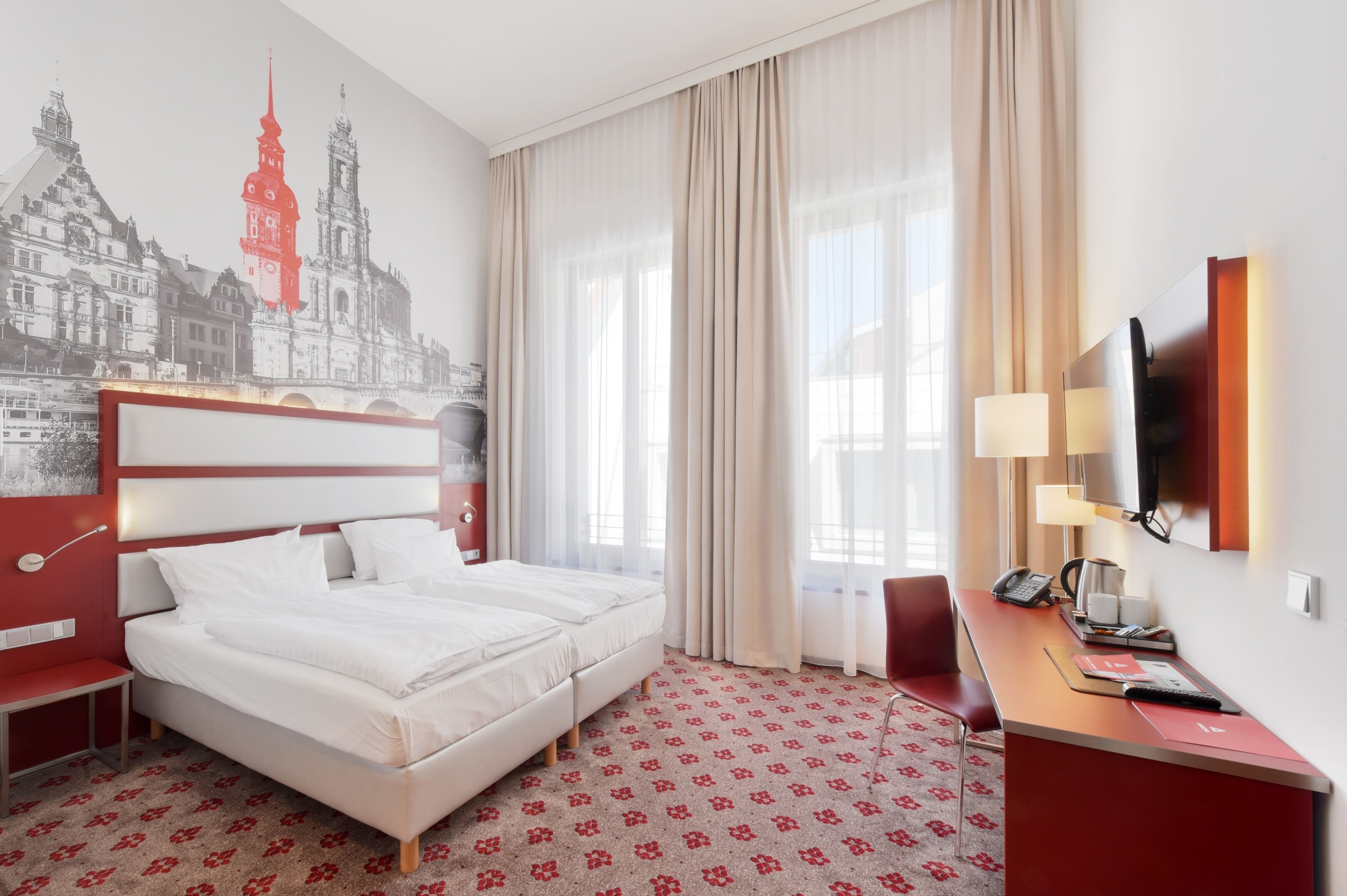 A hotel room with a bed, a desk with a telephone, a lamp, and a wall-mounted TV.