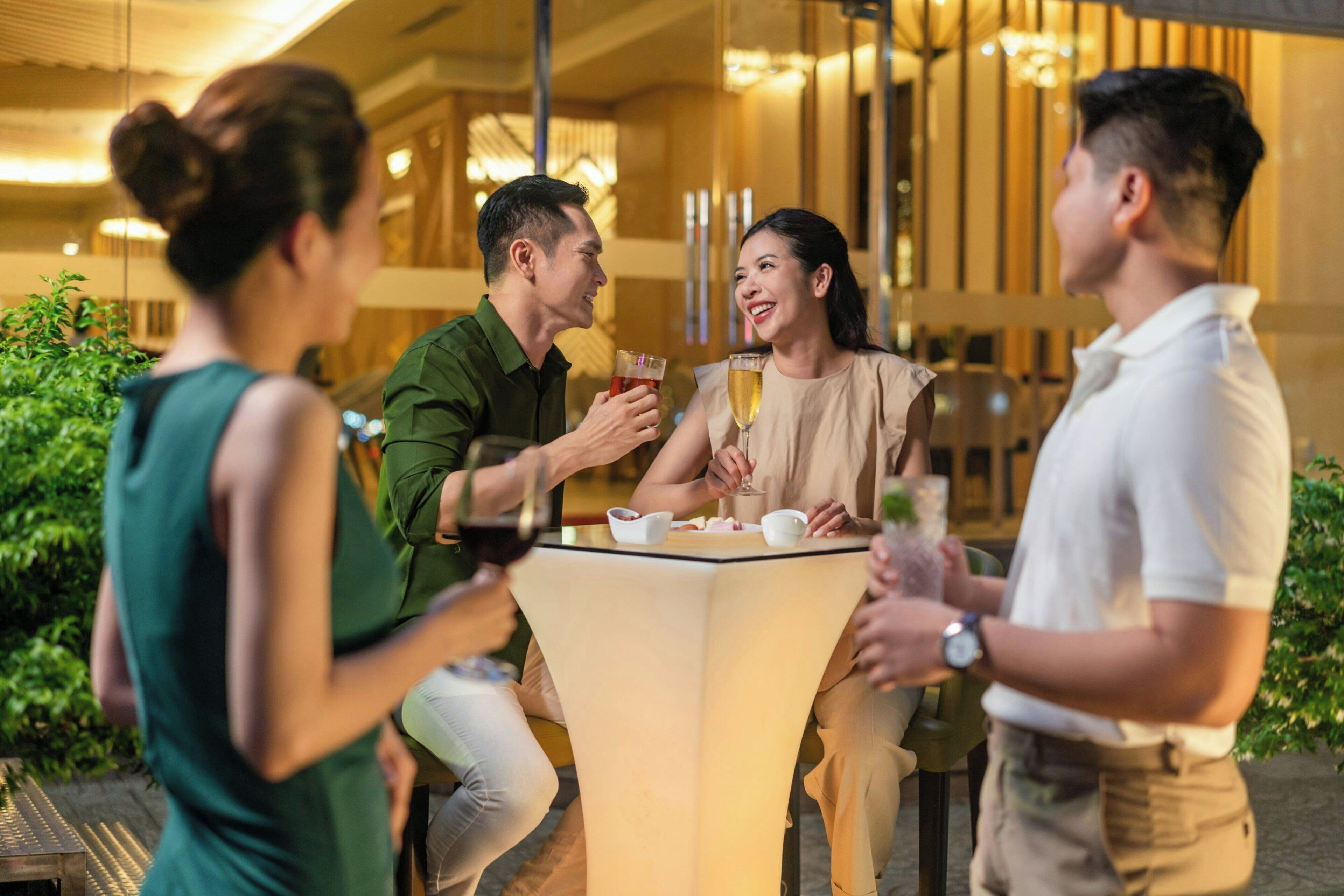 Un grupo de personas sentadas alrededor de una mesa con bebidas, en un ambiente interior cálidamente iluminado con plantas y decoración moderna.
