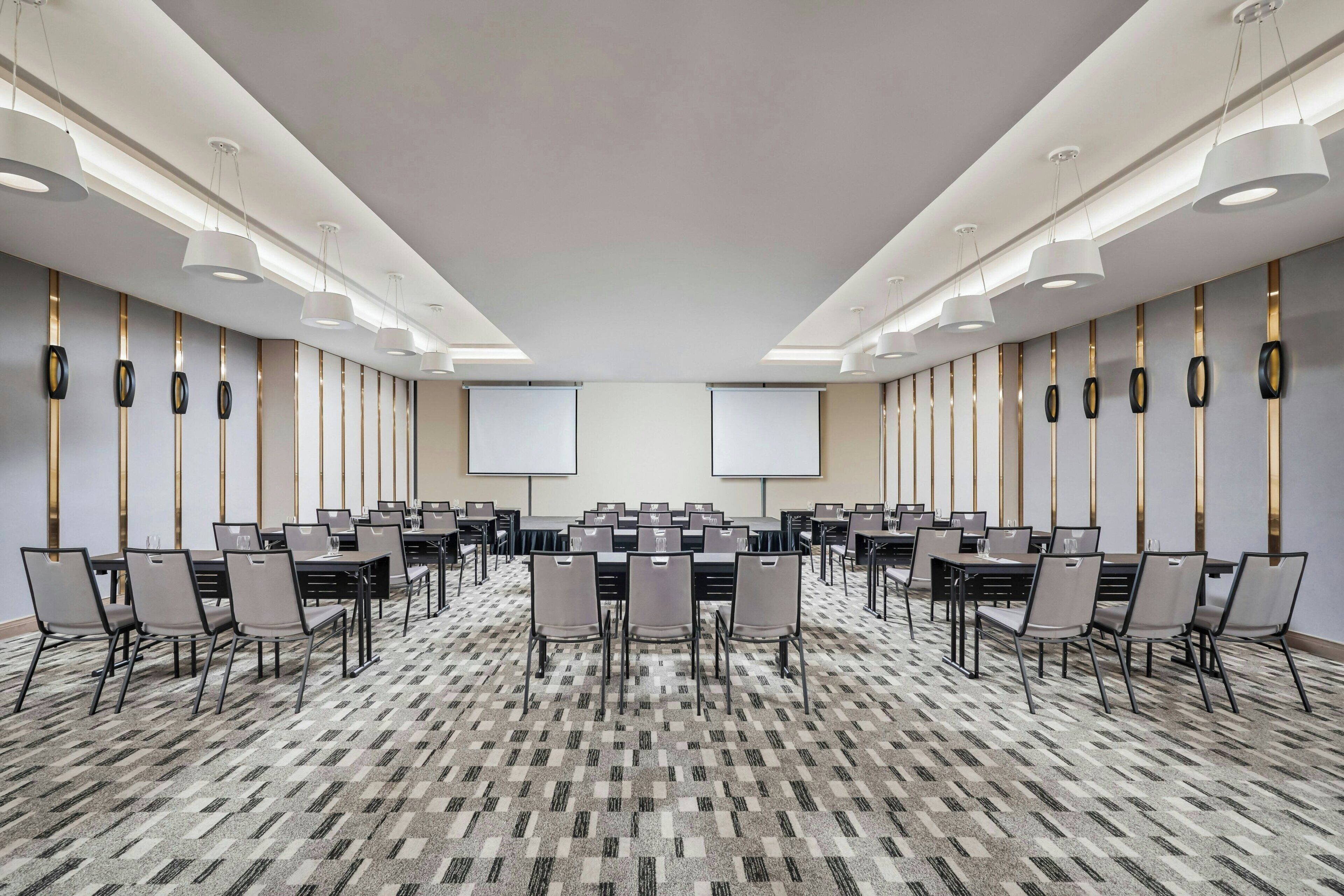 Un salón de conferencias con varias mesas y sillas, pantallas grandes y luminarias modernas.