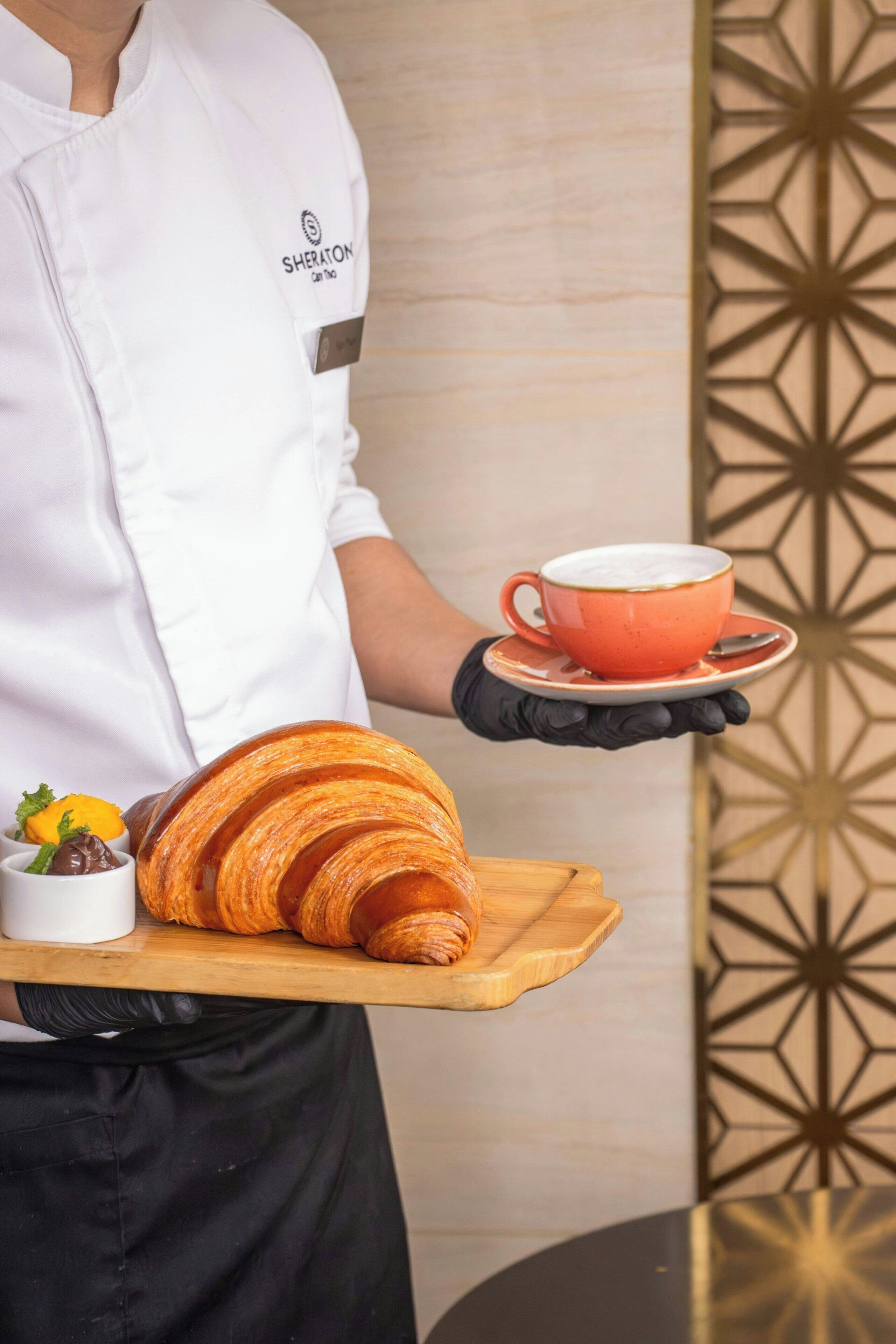 Una persona con uniforme blanco sostiene una bandeja con un croissant, una taza de café y dos pequeños recipientes de chocolate y mermelada.