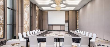 A conference room with a long table, white chairs, a projector screen, and a chandelier.