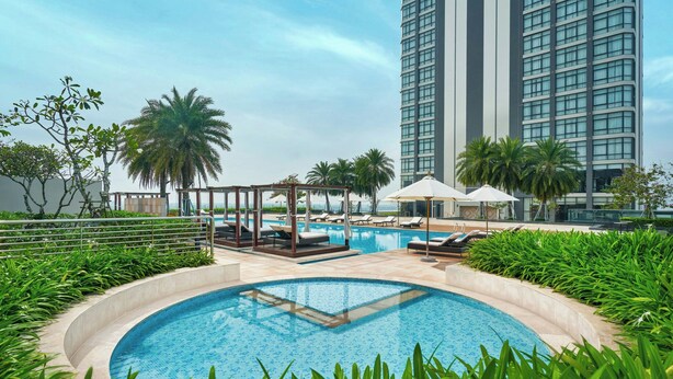 A pool area with lounge chairs and umbrellas, surrounded by greenery and a tall building in the background.