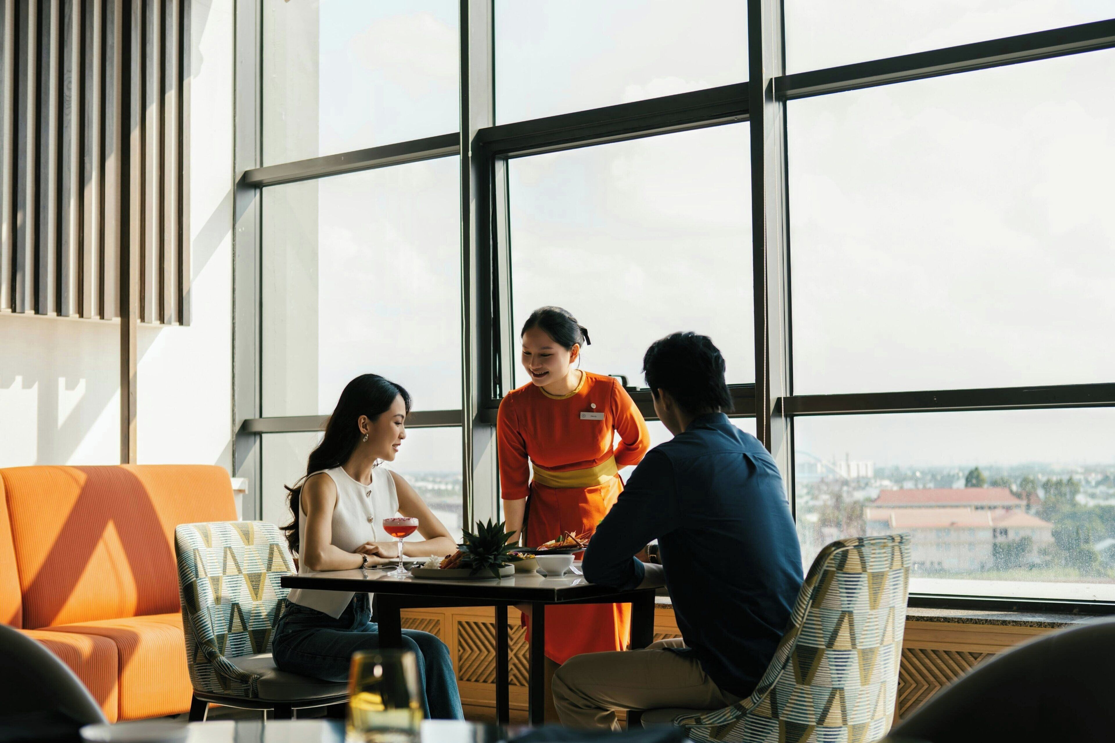 A dining area with a large window, a table with a person serving food, and a view of the cityscape.