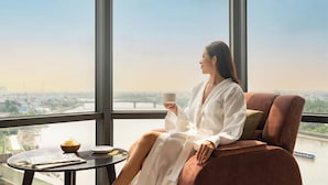 A person sitting on a cushioned chair by a window with a view of a cityscape and a river.