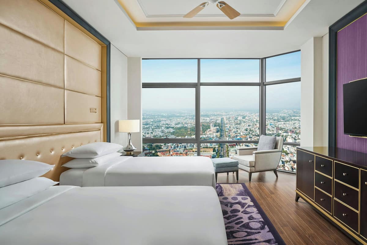 Habitación de hotel con dos camas, vistas a la ciudad, ventilador de techo y televisión en la pared.