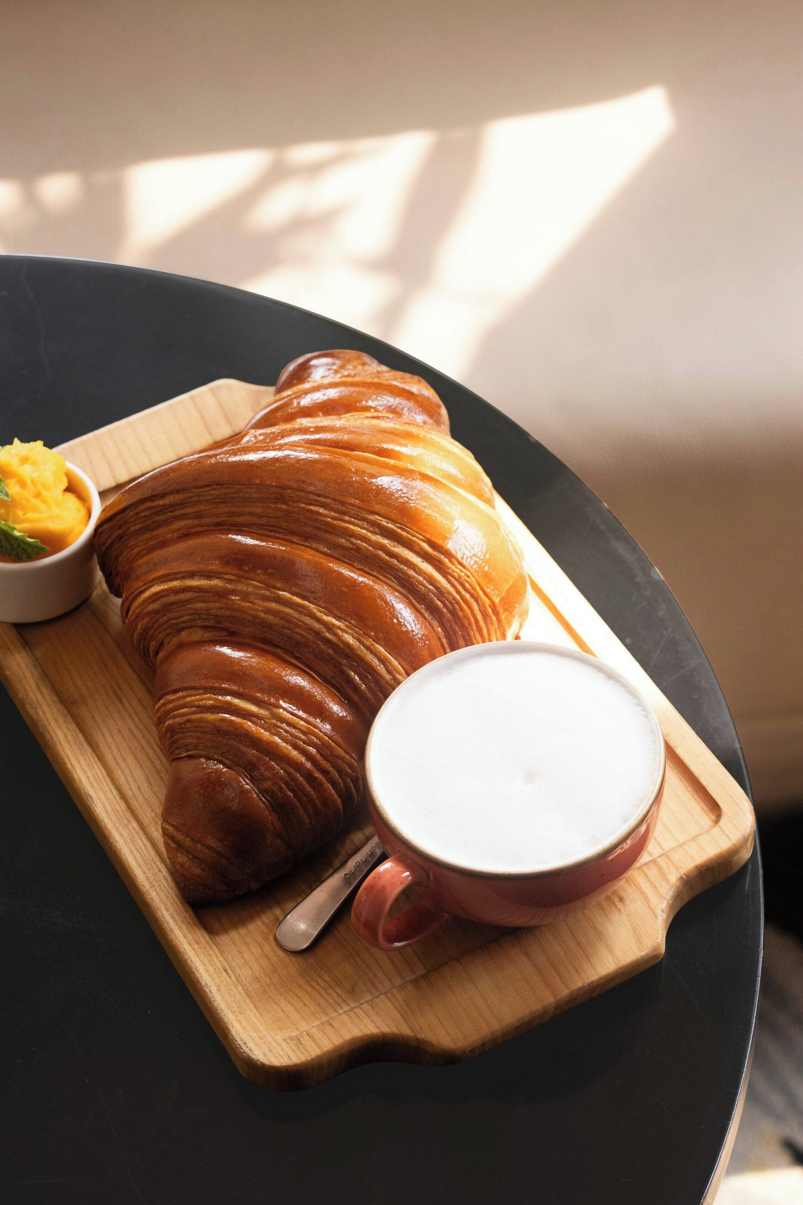 A croissant on a wooden board with a cup of coffee and a small bowl of yellow spread.