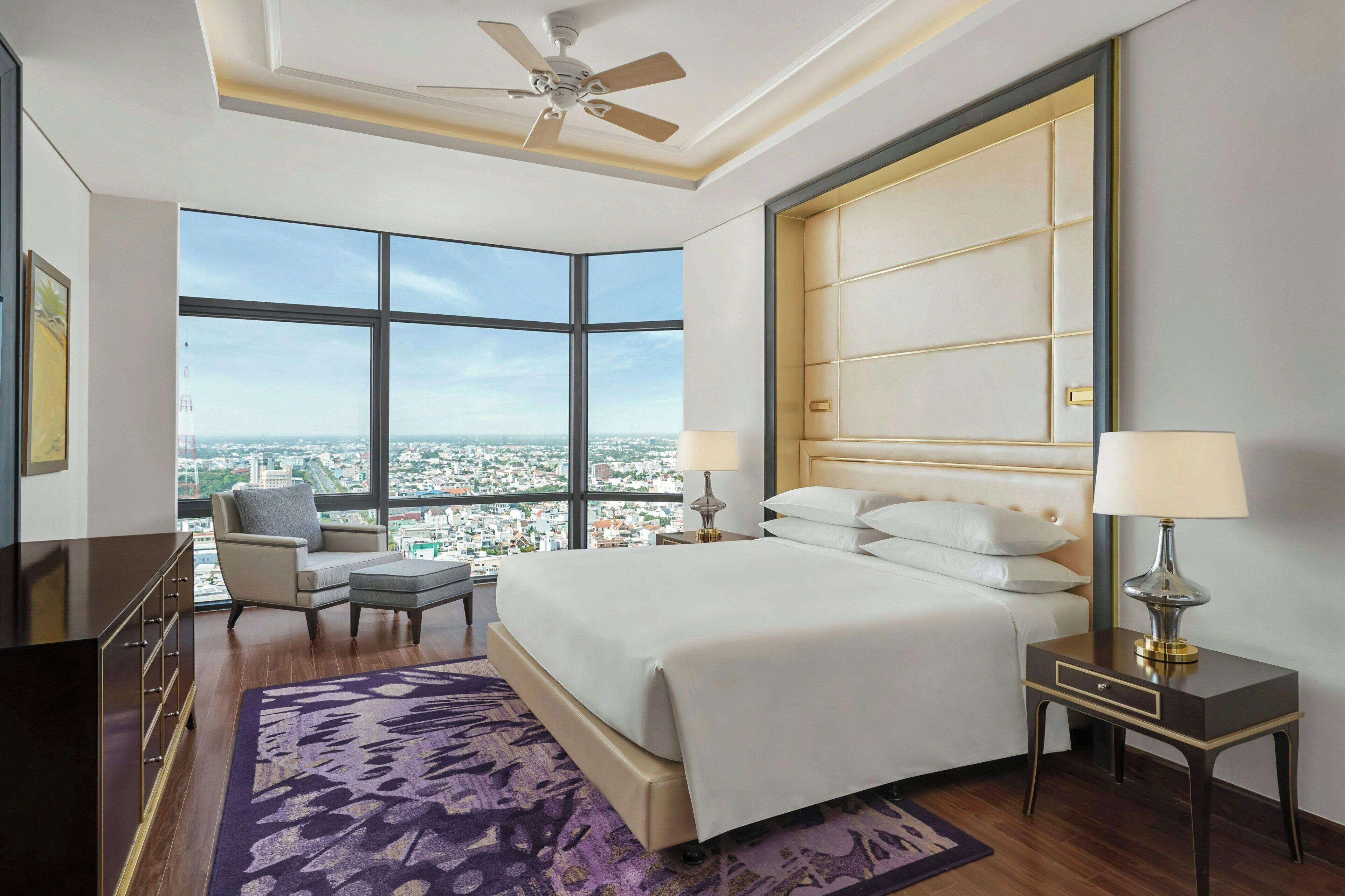 A hotel room with a large bed, two bedside tables with lamps, a chair, a dresser, and a view of a cityscape through large windows.