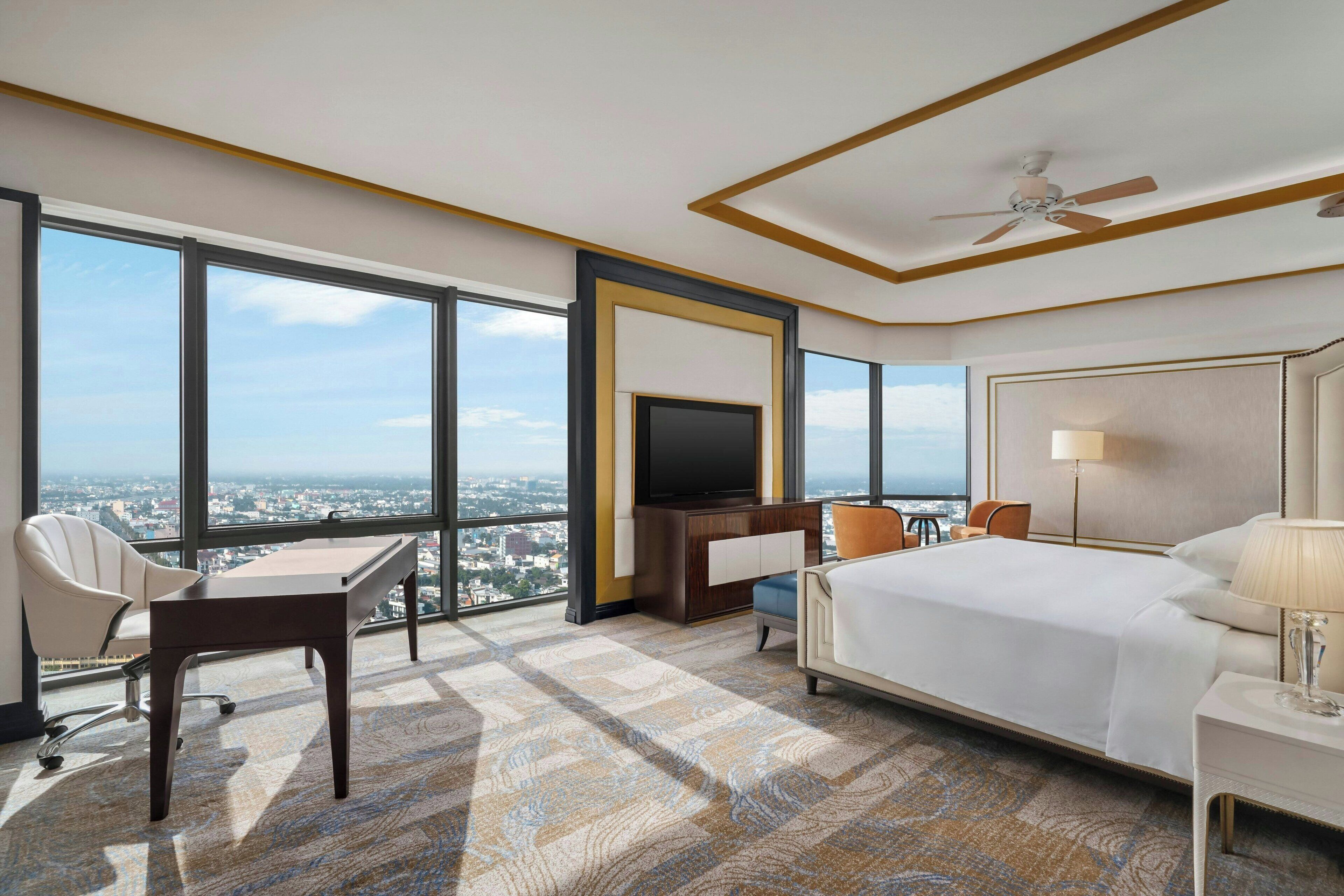 A hotel room with a large bed, a desk, a chair, a TV, and a view of the city.