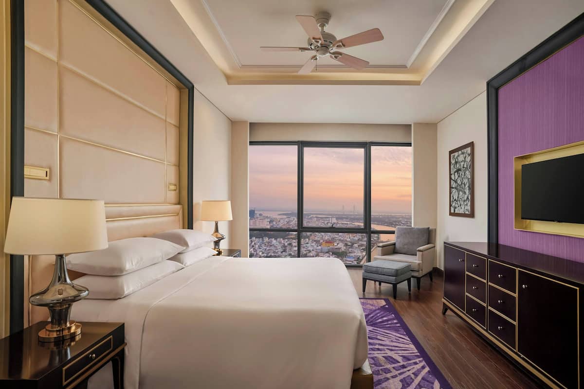 Habitación de hotel con una cama grande, dos mesitas de noche con lámparas, un ventilador de techo, televisión, una silla y vistas a la ciudad a través de amplias ventanas.
