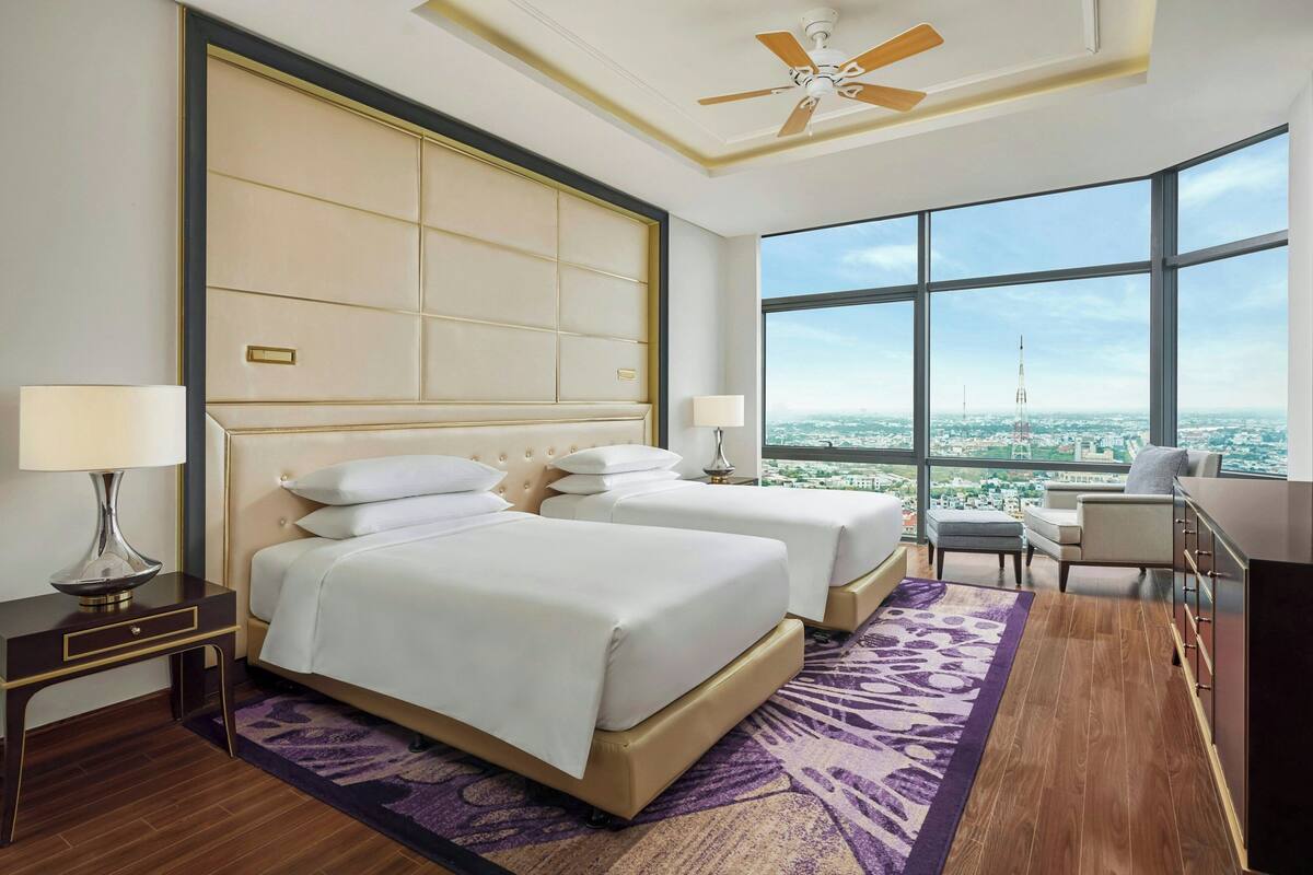Habitación de hotel con dos camas, un ventanal amplio con vistas a la ciudad, un ventilador de techo y un escritorio con lámpara.