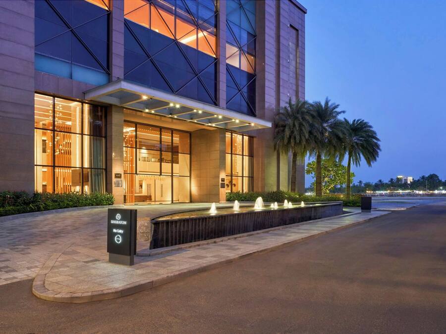 Een moderne hotelentree met een goed verlichte gevel, een waterpartij ervoor en palmbomen in de buurt.