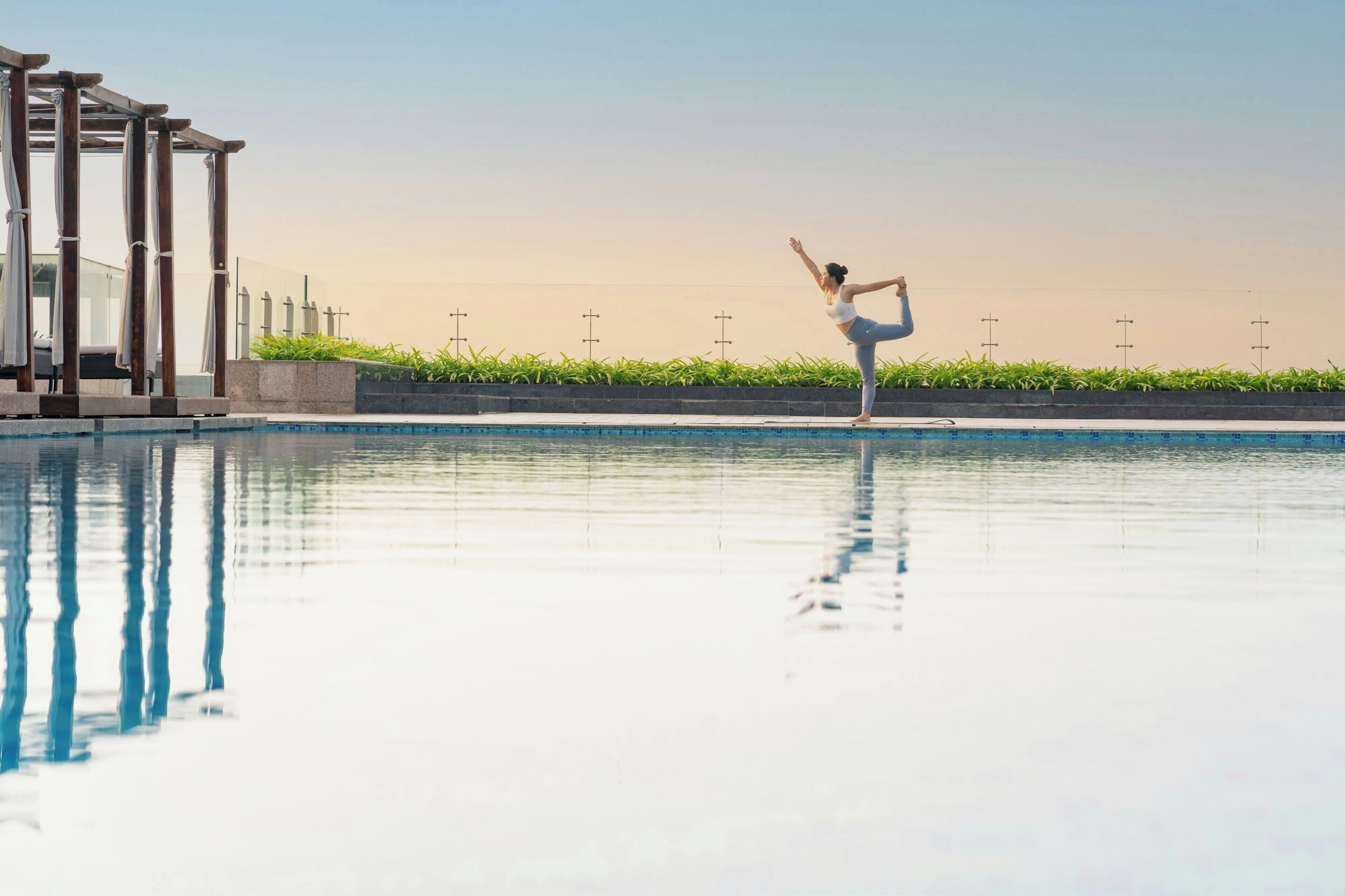 Una persona realizando una postura de yoga junto a una piscina con una pérgola y follaje al fondo.
