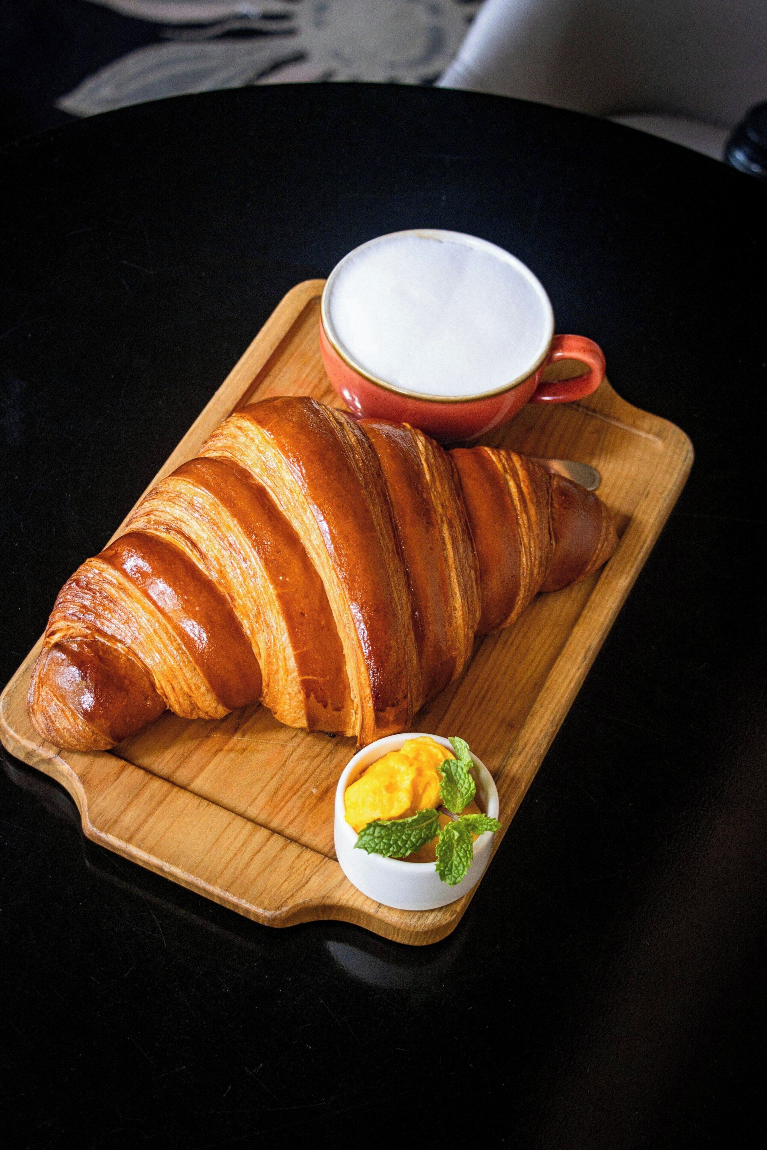A croissant on a wooden board with a small bowl of fruit and a cup of coffee.