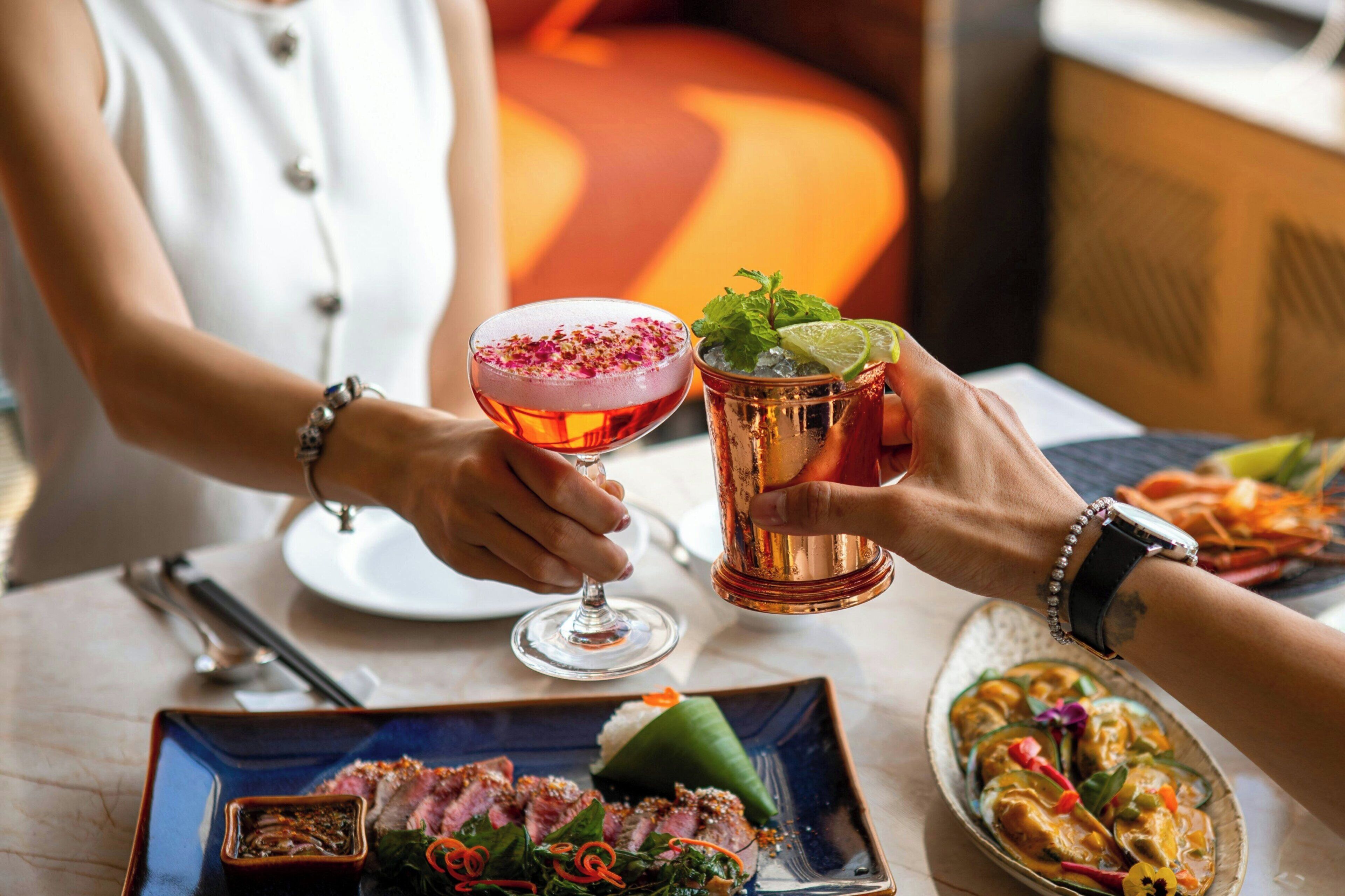 A dining table with a plate of sliced meat, a cocktail, and a glass of a beverage with a garnish.
