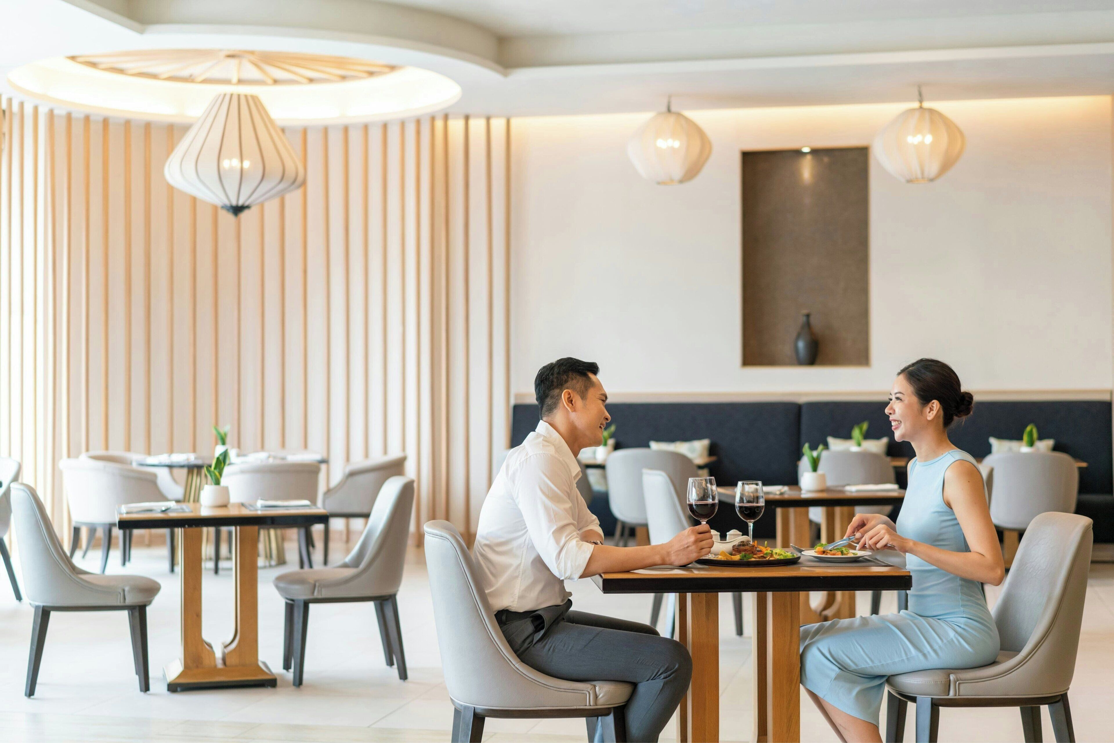 A modern dining area with a man and a woman seated at a table, both engaged in conversation. The room features contemporary furniture, pendant lighting, and a neutral color palette.